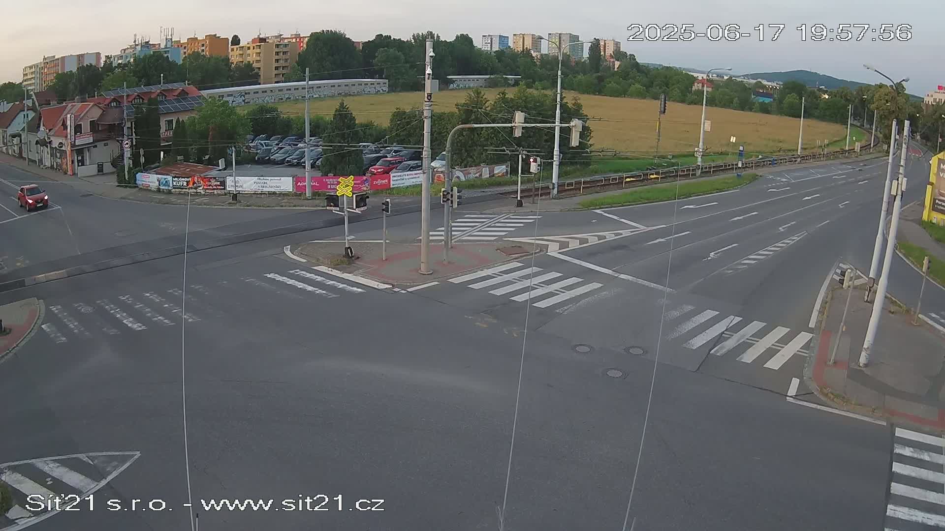 A high-angle, long shot of a multi-lane road intersection with crosswalks, traffic signals, and a few cars; it is daytime with partly cloudy skies.