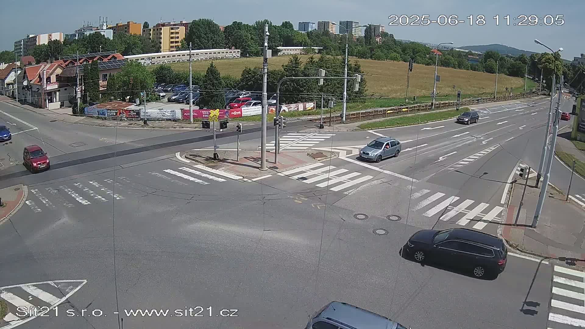 A sunny day shows a multi-lane intersection with several cars and crosswalks, buildings and a grassy field in the background.