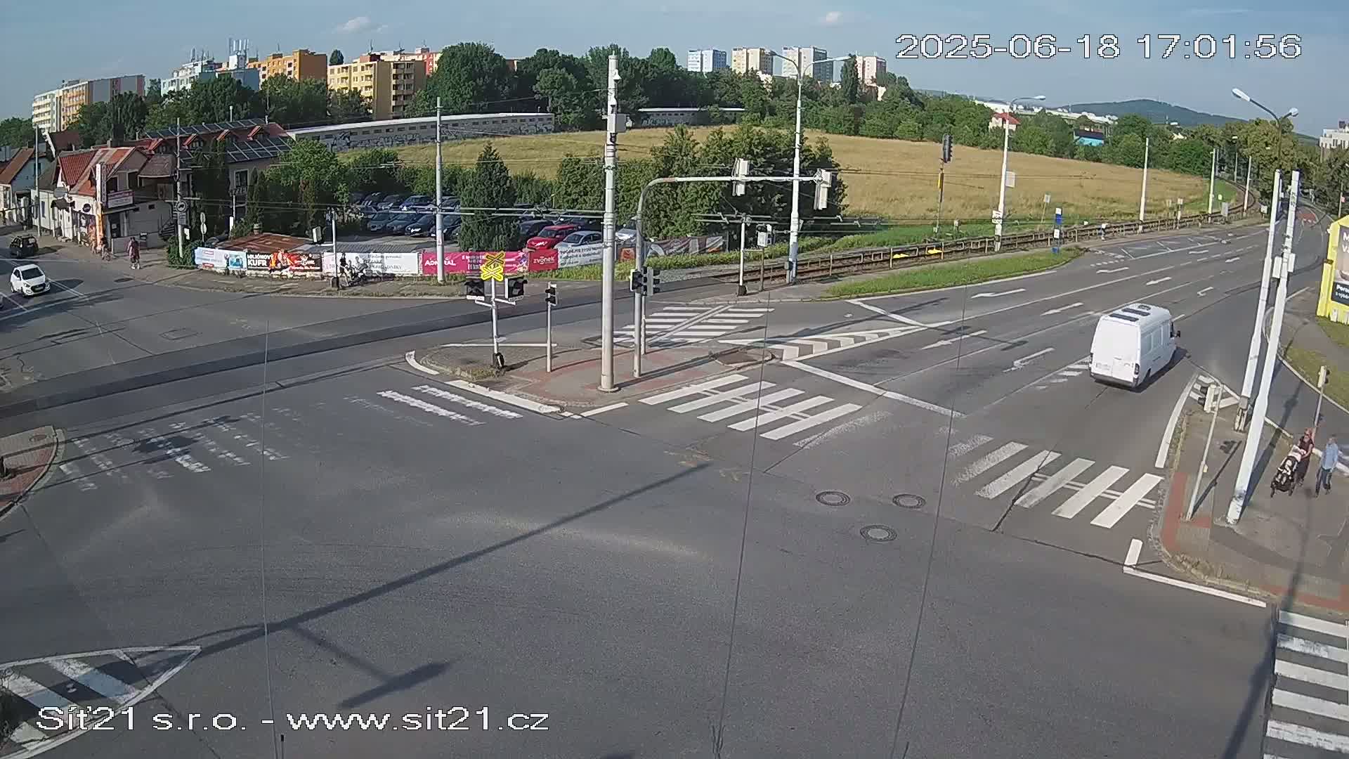 A sunny day shows a multi-lane intersection with crosswalks, traffic signals, and several vehicles, including a white van, with pedestrians near the curb.