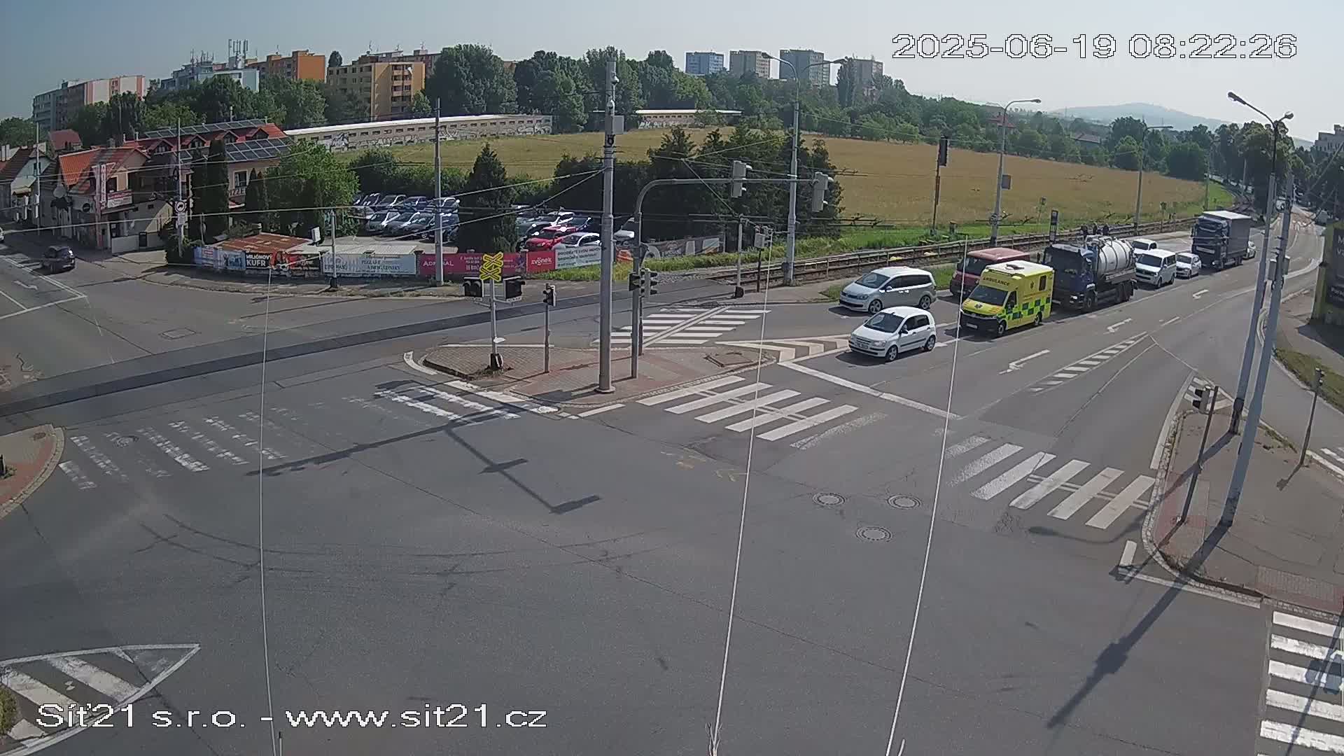 A sunny day shows a multi-lane intersection with several cars and an ambulance stopped at a crosswalk, while other vehicles are driving past a parking lot and buildings in the background.