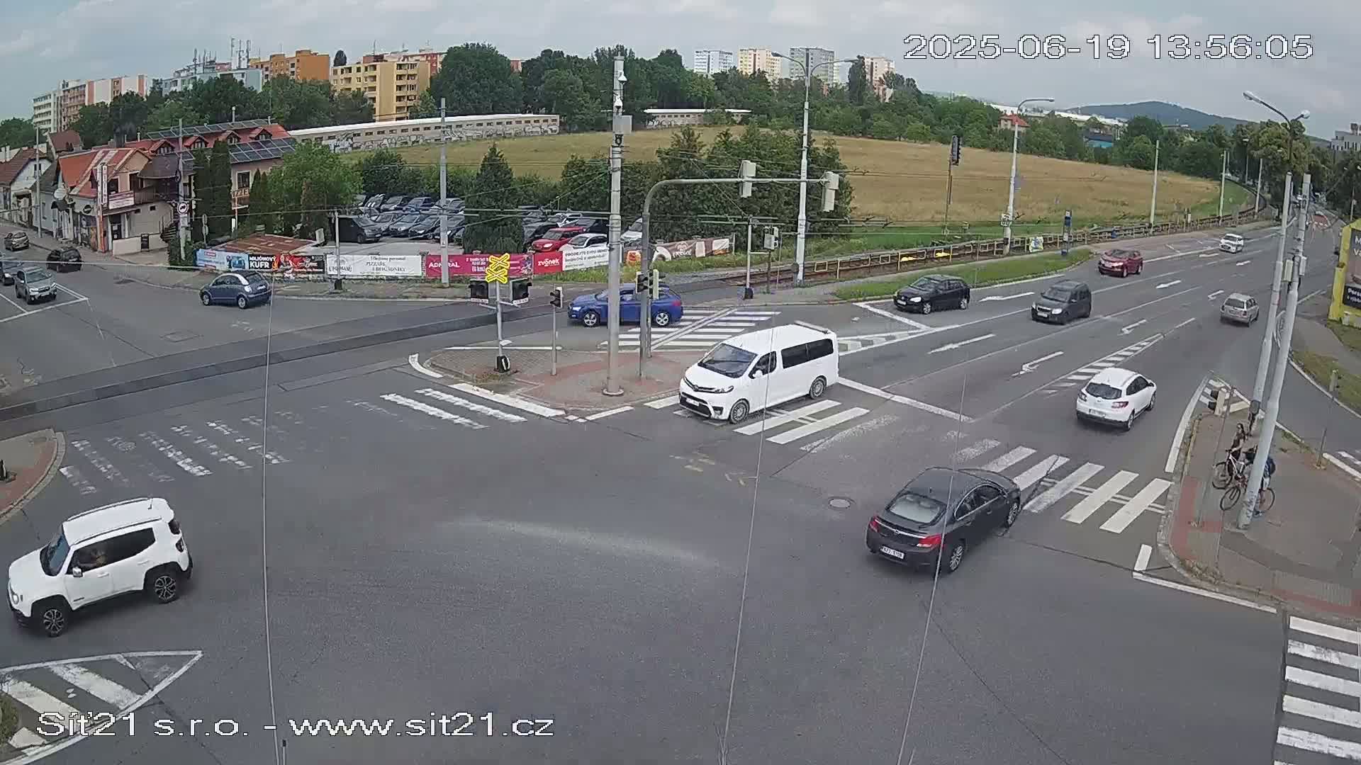 A high-angle view of a multi-lane intersection with several cars and a van at a crosswalk under partly cloudy skies.