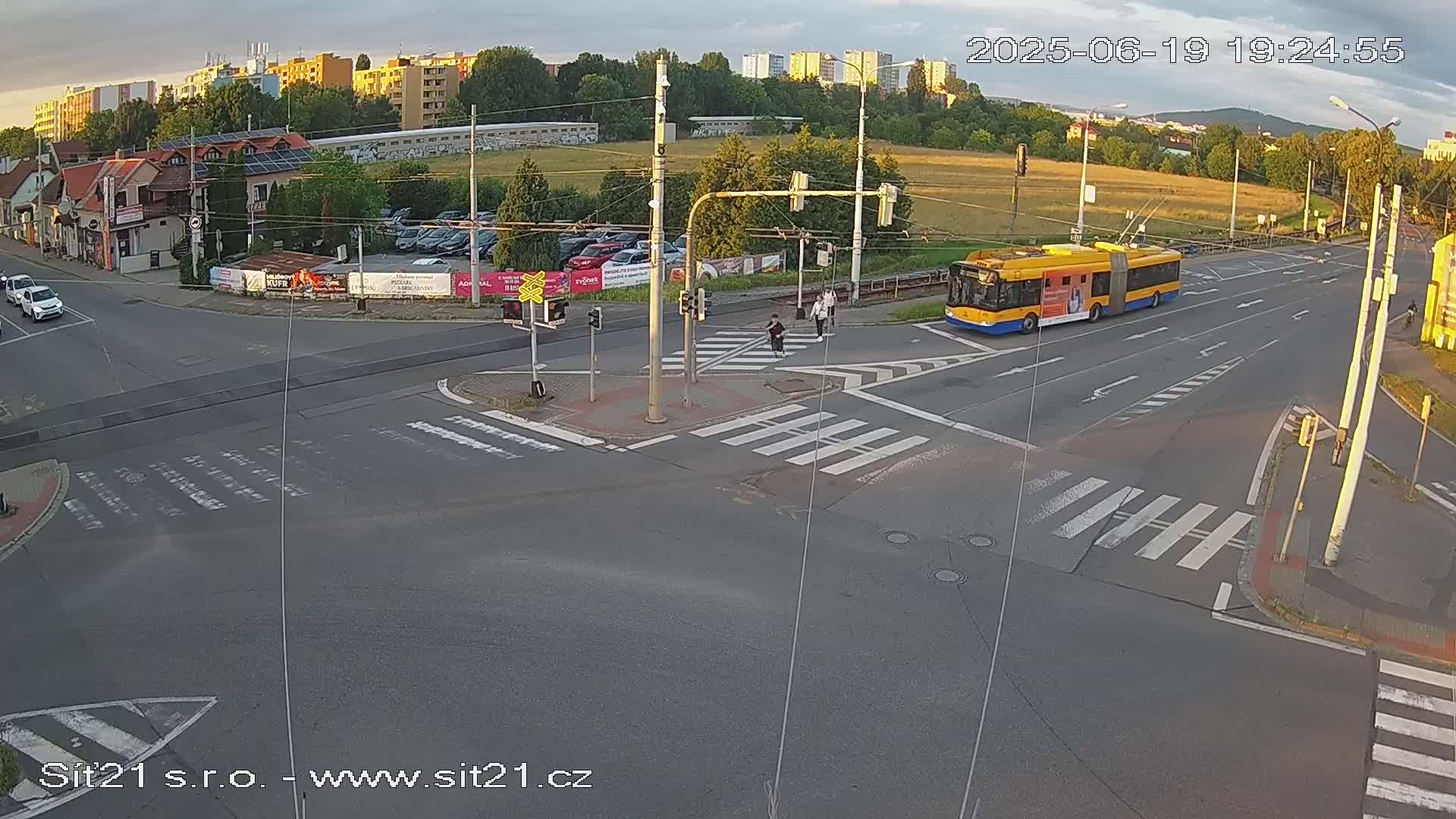 A yellow bus drives past a crosswalk where pedestrians are walking, at a multi-lane intersection under a partly cloudy sunset sky.