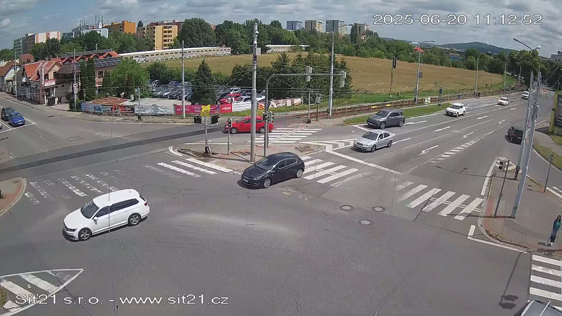 A sunny day shows several cars navigating a multi-lane intersection with pedestrian crosswalks, surrounded by buildings and green spaces.