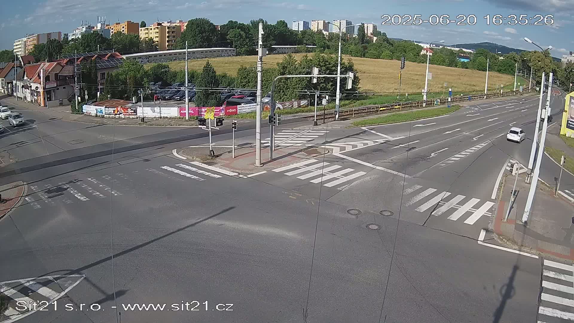 A mostly sunny overhead view of a multi-lane road intersection with crosswalks and traffic signals, featuring several cars and buildings in the background.