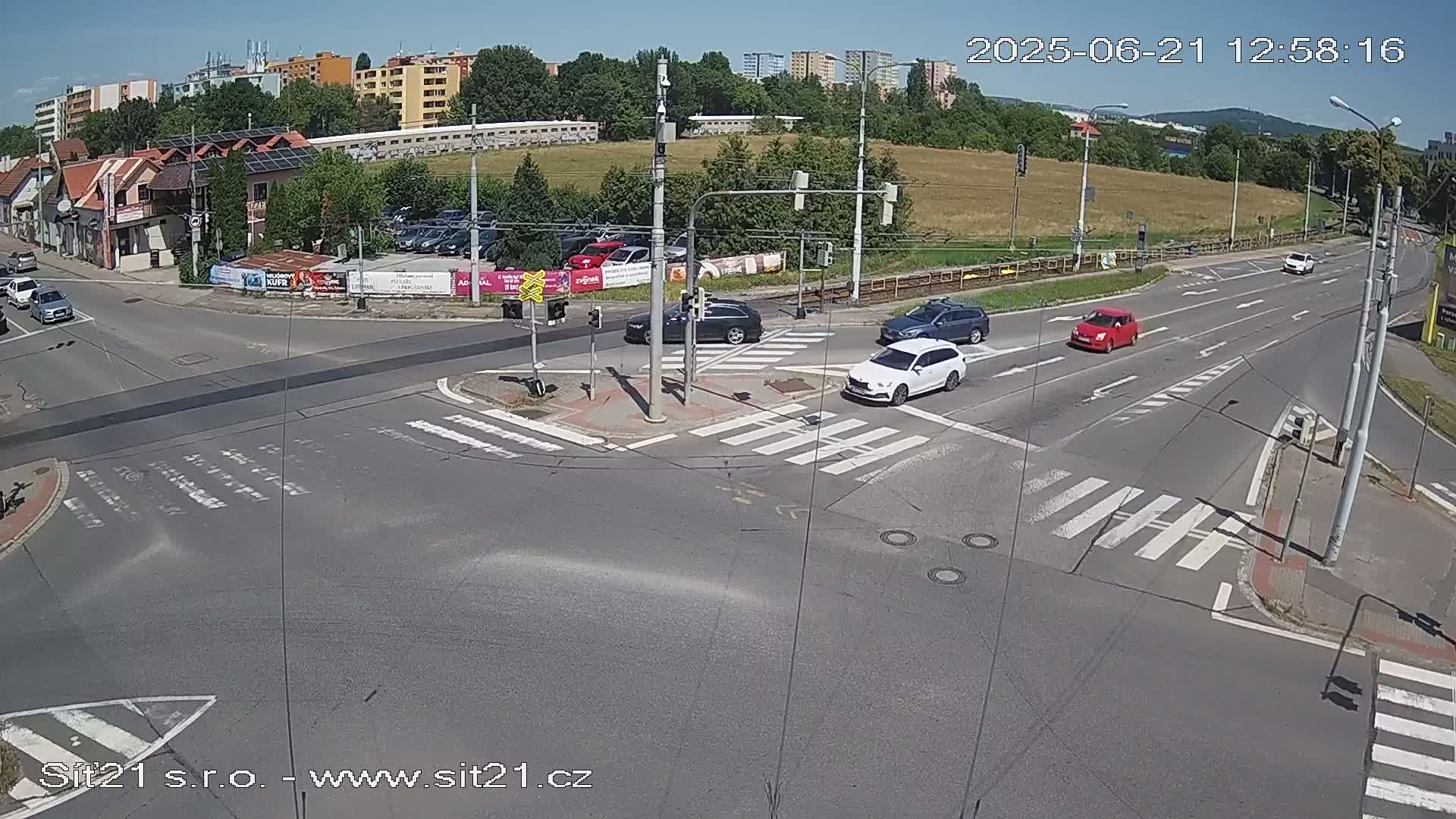 A sunny day shows several cars navigating a multi-lane intersection with pedestrian crosswalks, surrounded by buildings and a grassy area.