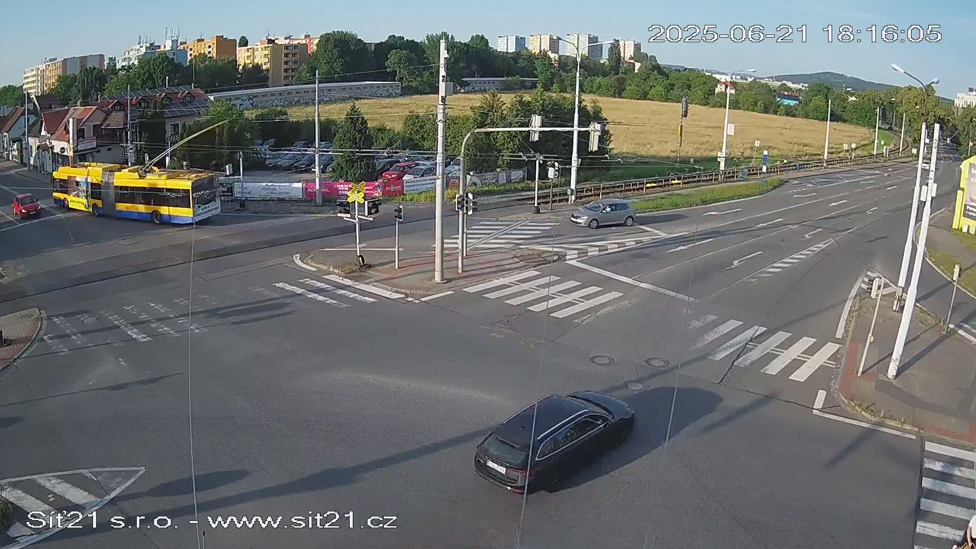 A sunny day shows a street intersection with a trolley bus, several cars, and crosswalks.