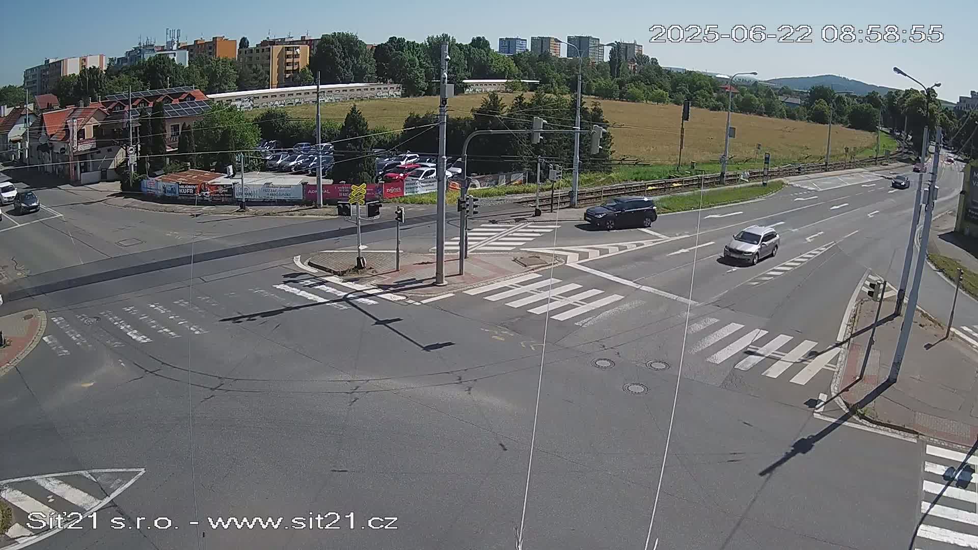 A sunny day shows a multi-lane road intersection with crosswalks, several cars, and buildings in the background.