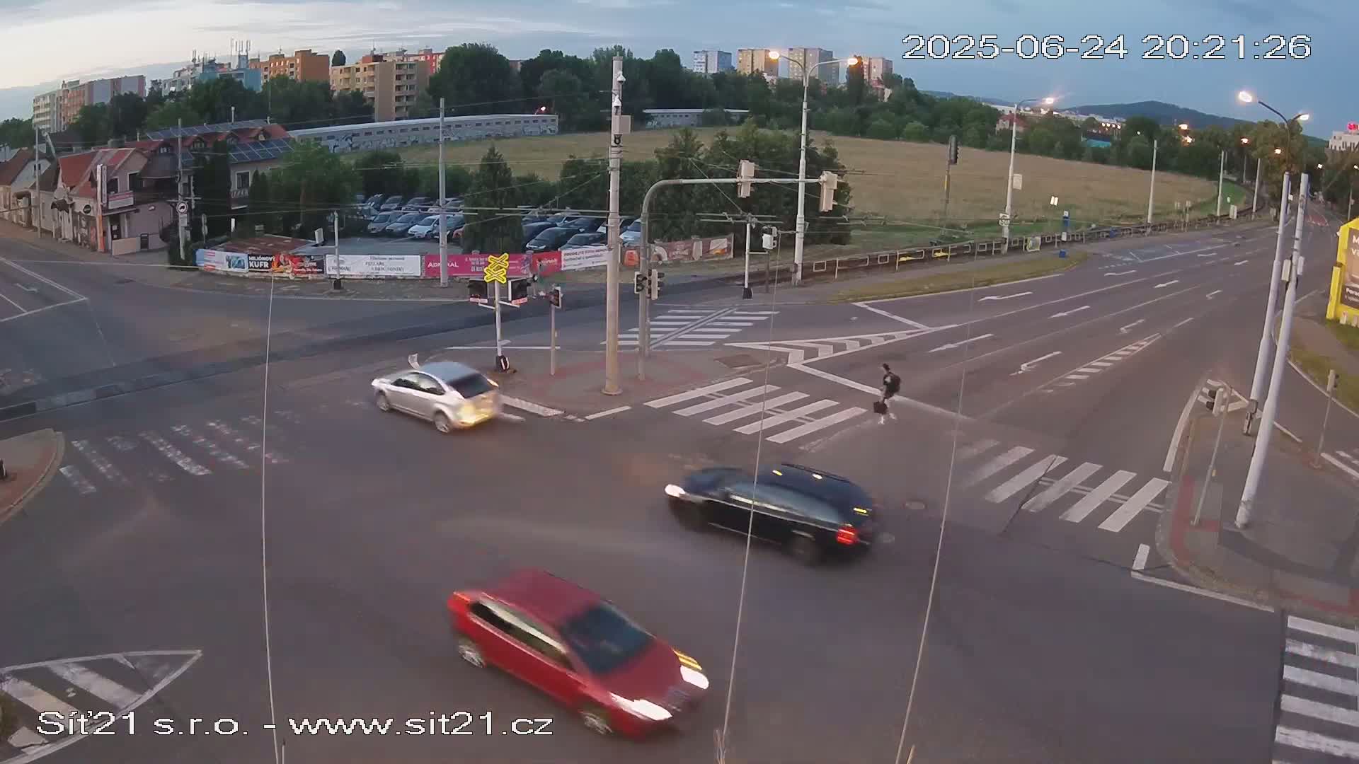 A twilight view of a multi-lane road intersection with several cars, a pedestrian crossing, and buildings in the background.