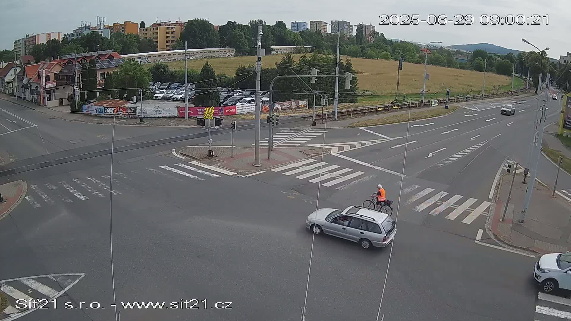 A mostly empty road intersection with a silver station wagon and a bicyclist crossing, under overcast skies.