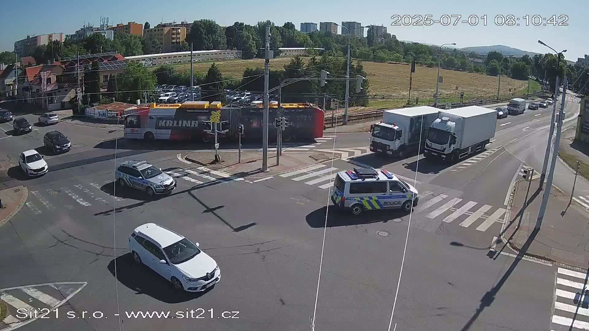 A sunny day shows a busy intersection with several cars, two trucks, a bus, and a police van, all at a crossroads.