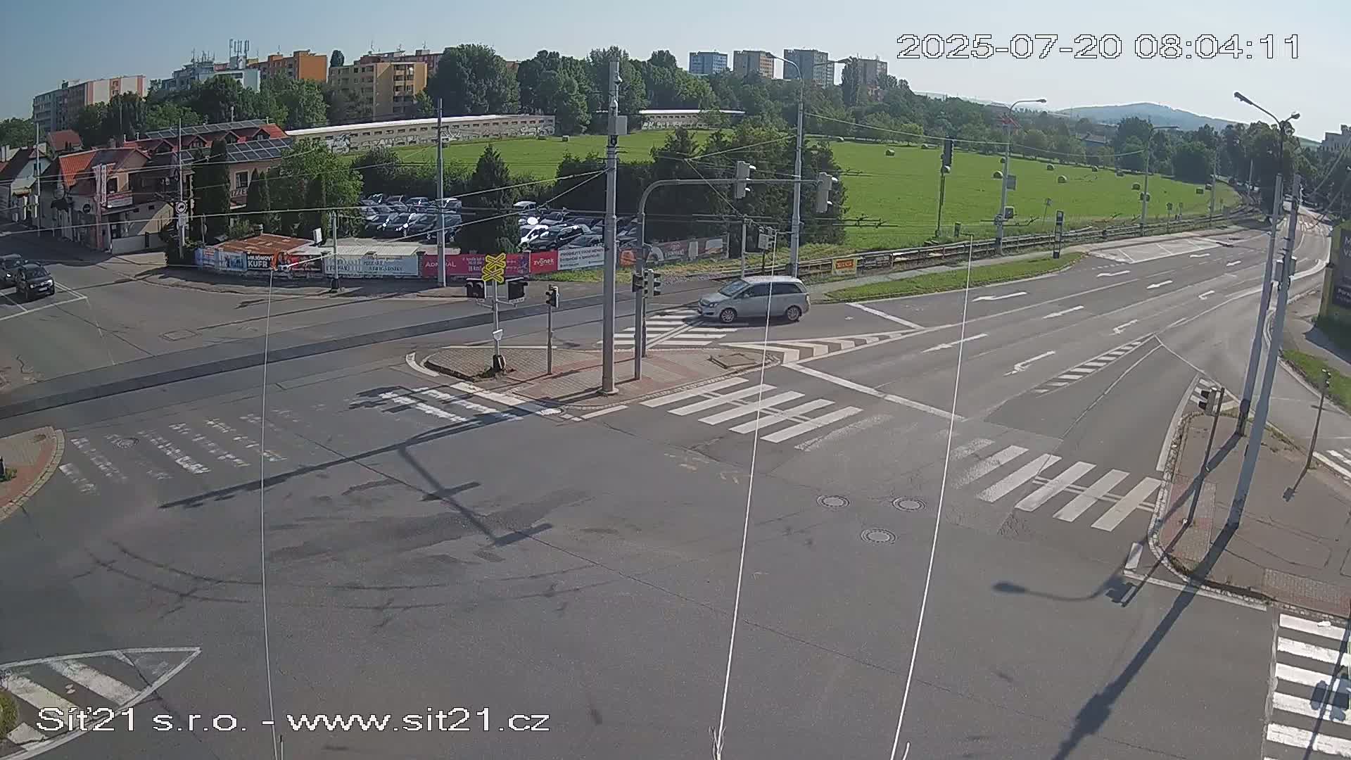 A sunny day shows a multi-lane road intersection with crosswalks, traffic lights, and a few vehicles, including one silver car, near apartment buildings and a grassy field.