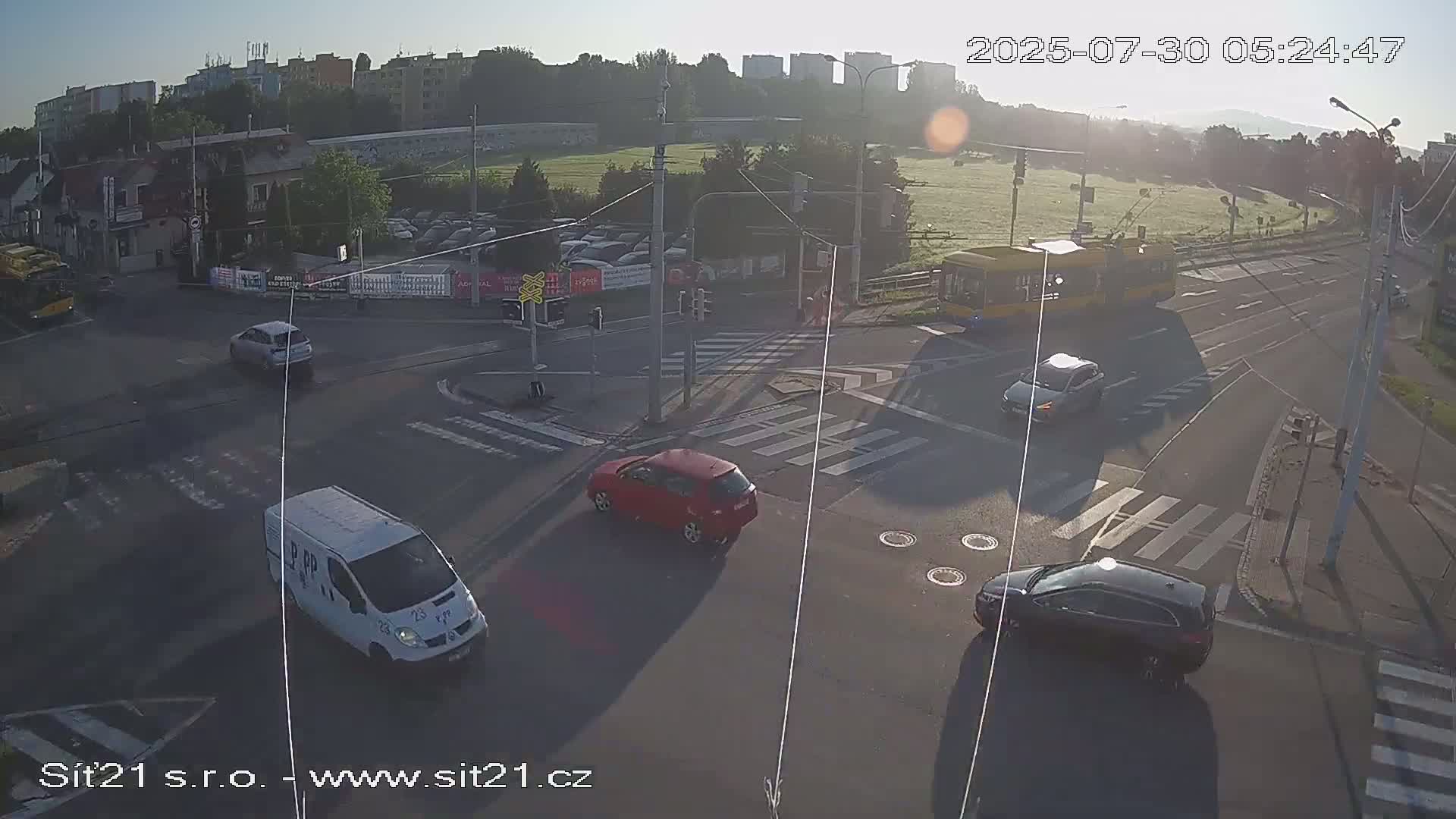 A sunny day shows a multi-lane road intersection with crosswalks, traffic lights, and a few vehicles, including one silver car, near apartment buildings and a grassy field.