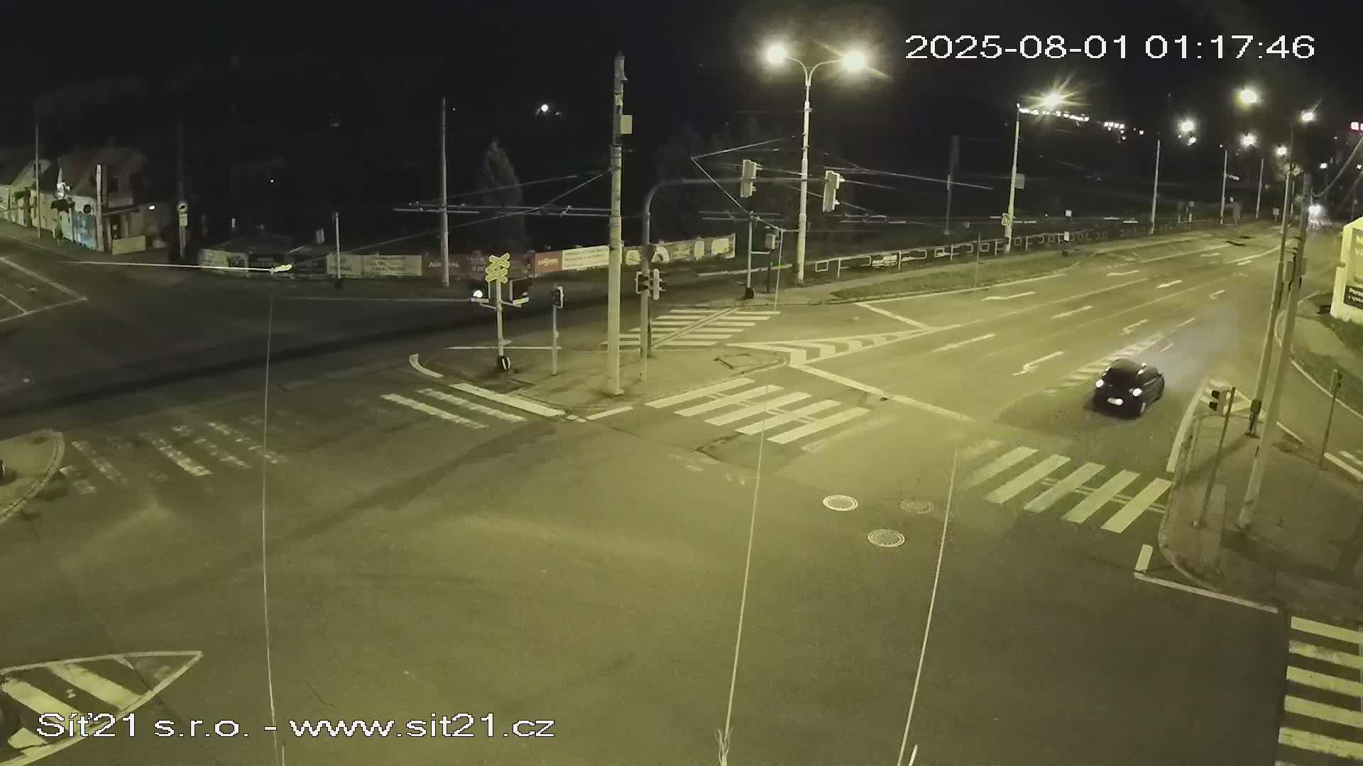A dark-colored car drives through a well-lit intersection at night.