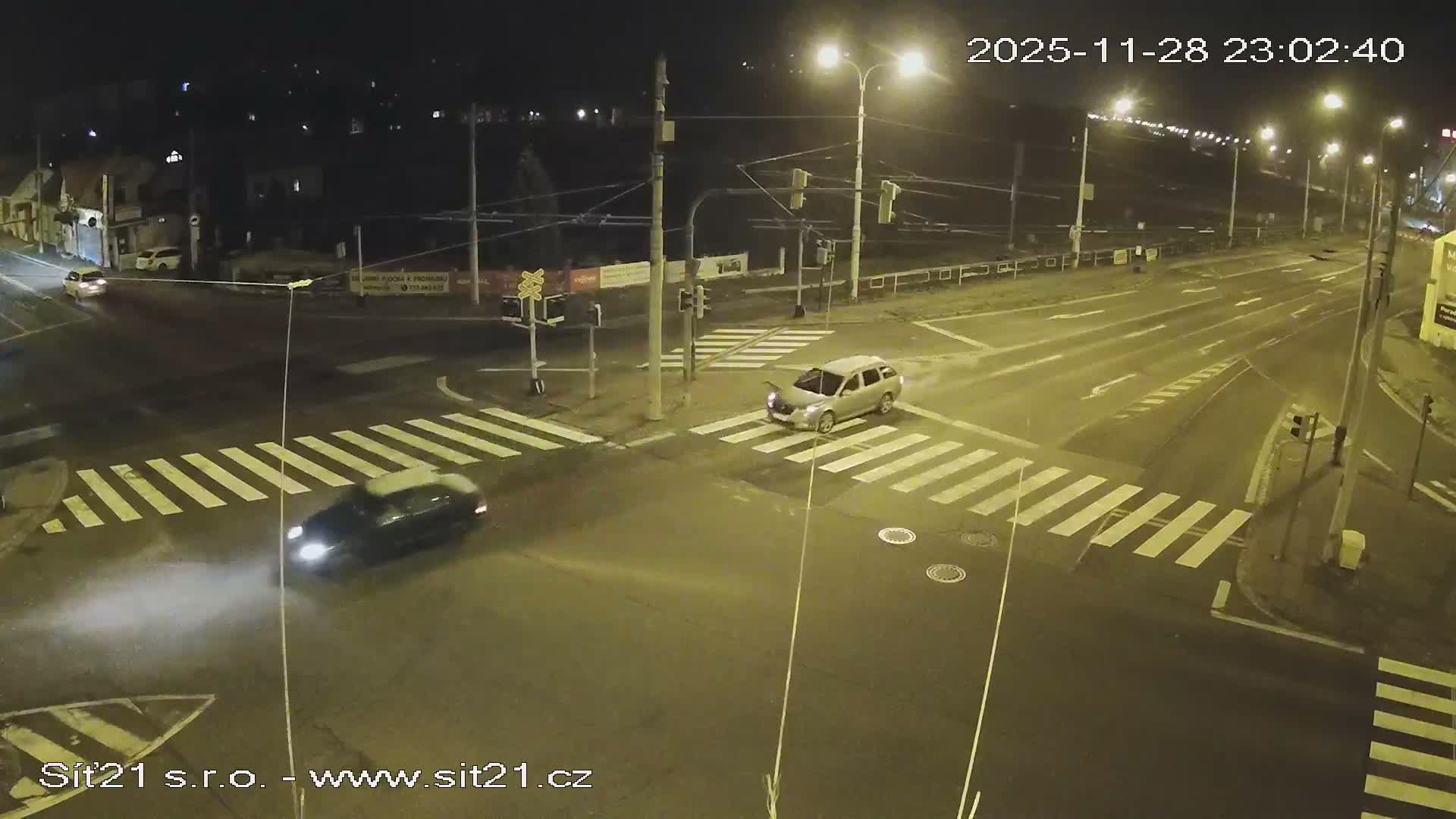 An elevated night-time view captures an urban intersection with wet roads, showing a silver SUV stopped at a crosswalk with its hood open, and a dark car with bright headlights moving in the foreground with visible exhaust.