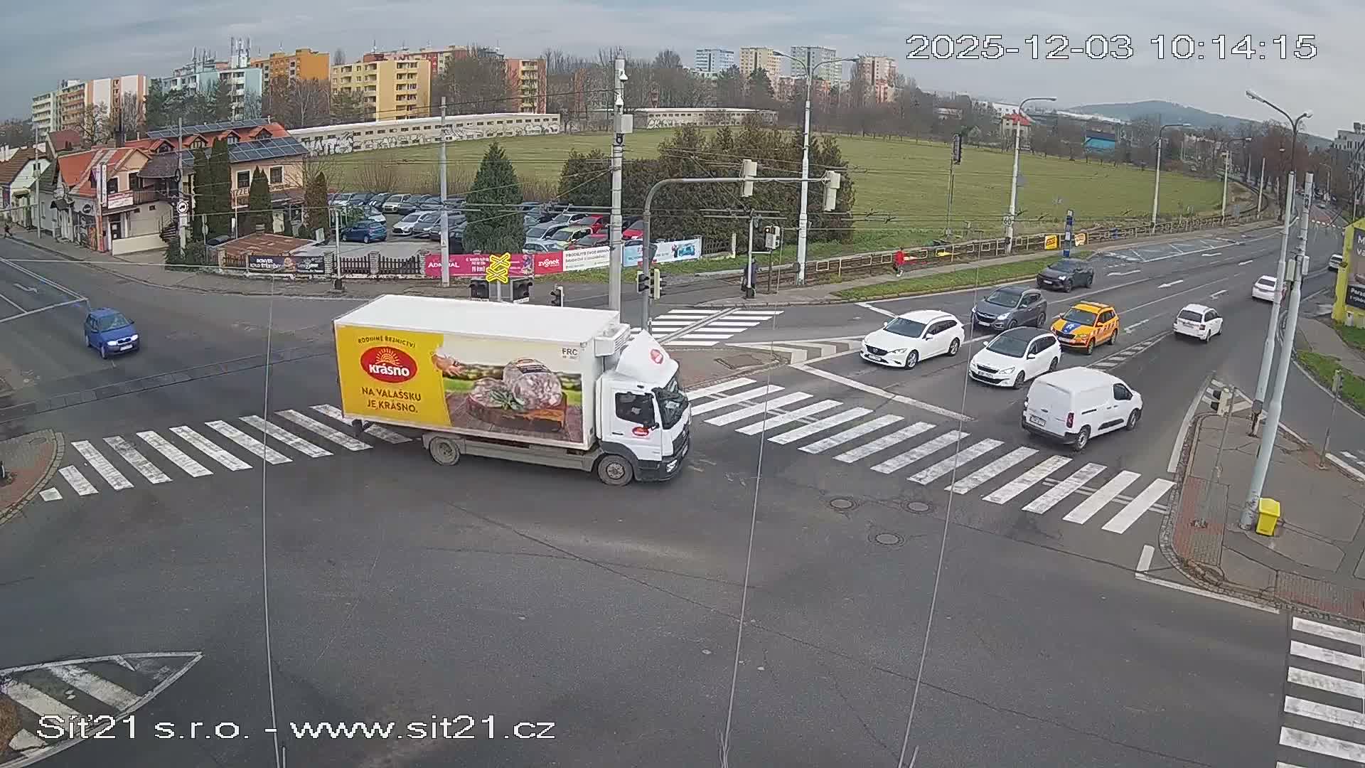 On a cloudy day, a white delivery truck with a food advertisement turns left at a bustling urban intersection, surrounded by several cars and residential buildings, with a large grassy area visible in the background.