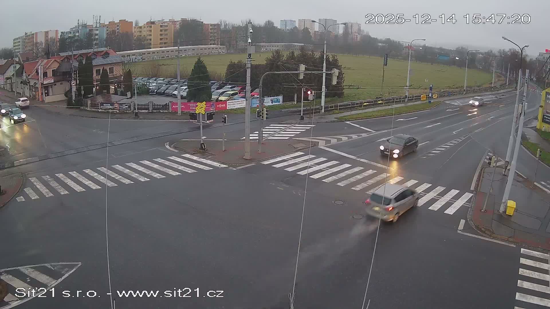 On a cloudy day, a white delivery truck with a food advertisement turns left at a bustling urban intersection, surrounded by several cars and residential buildings, with a large grassy area visible in the background.