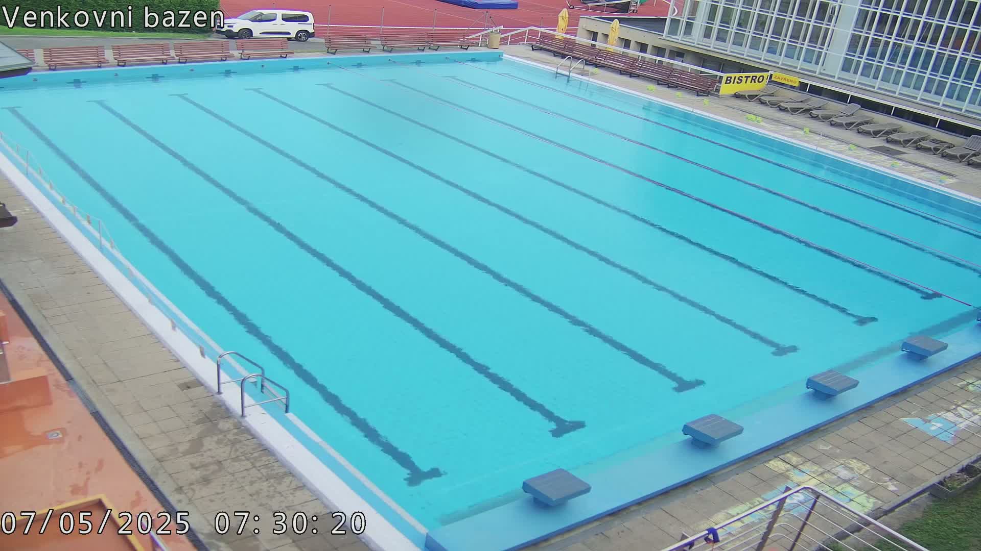 An outdoor swimming pool with several lanes is empty and mostly clear, under an overcast sky.