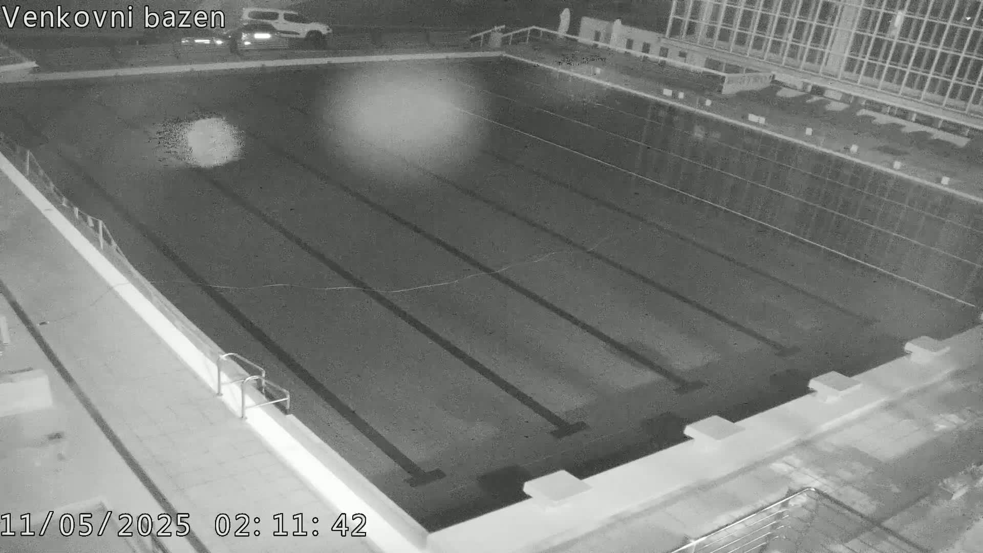 An empty outdoor swimming pool at night is viewed from above, showing lane dividers and a vehicle parked nearby.