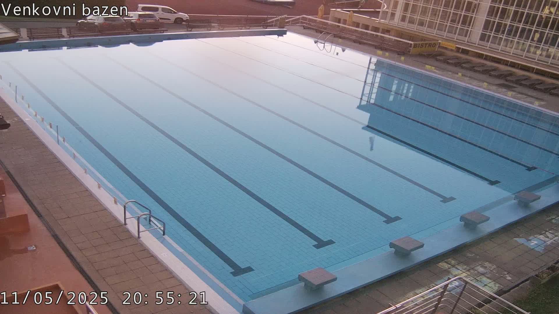 An empty, light-blue outdoor swimming pool with multiple lanes reflects the surrounding buildings under a clear sky.