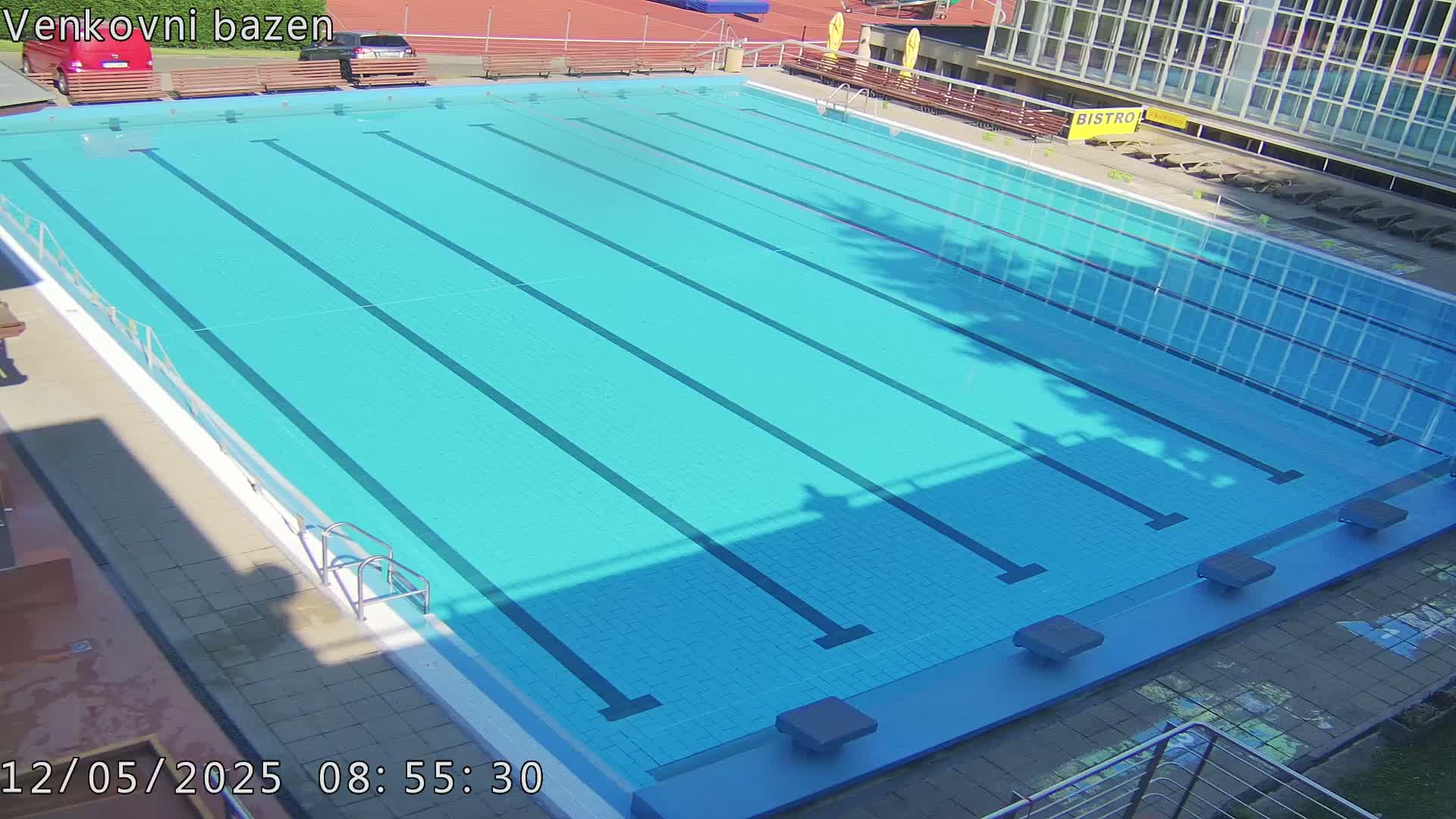 An outdoor, sunny swimming pool with multiple lanes is empty, surrounded by benches and a building.