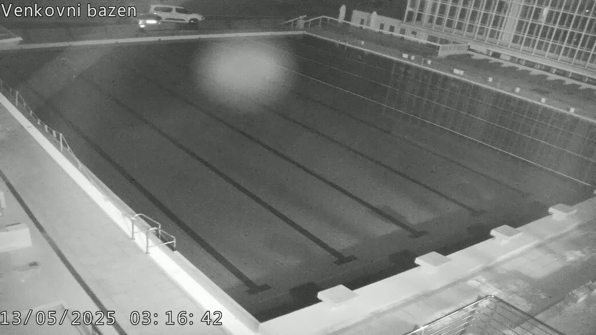 An empty, dark swimming pool is seen at night, with a vehicle visible in the distance.
