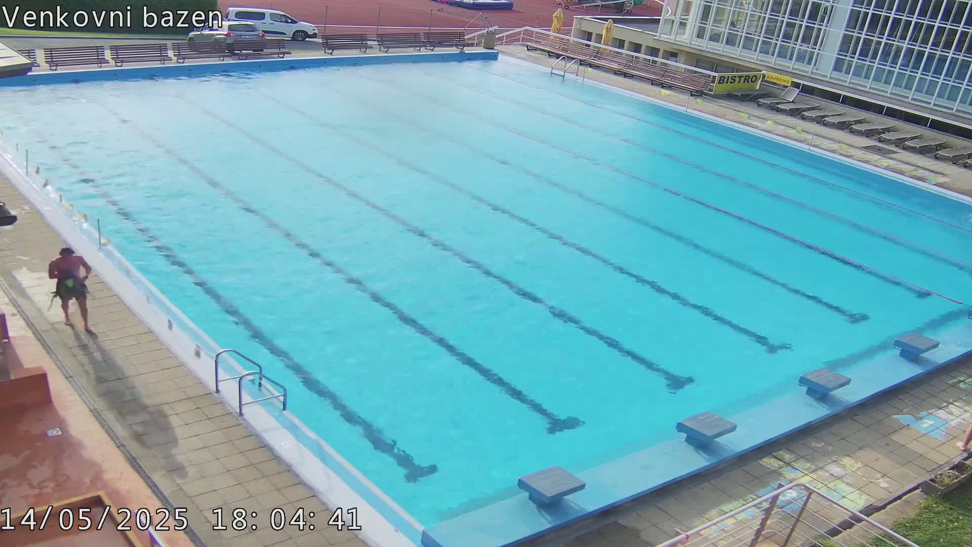 A person walks beside a large, light-blue outdoor swimming pool with several lanes.