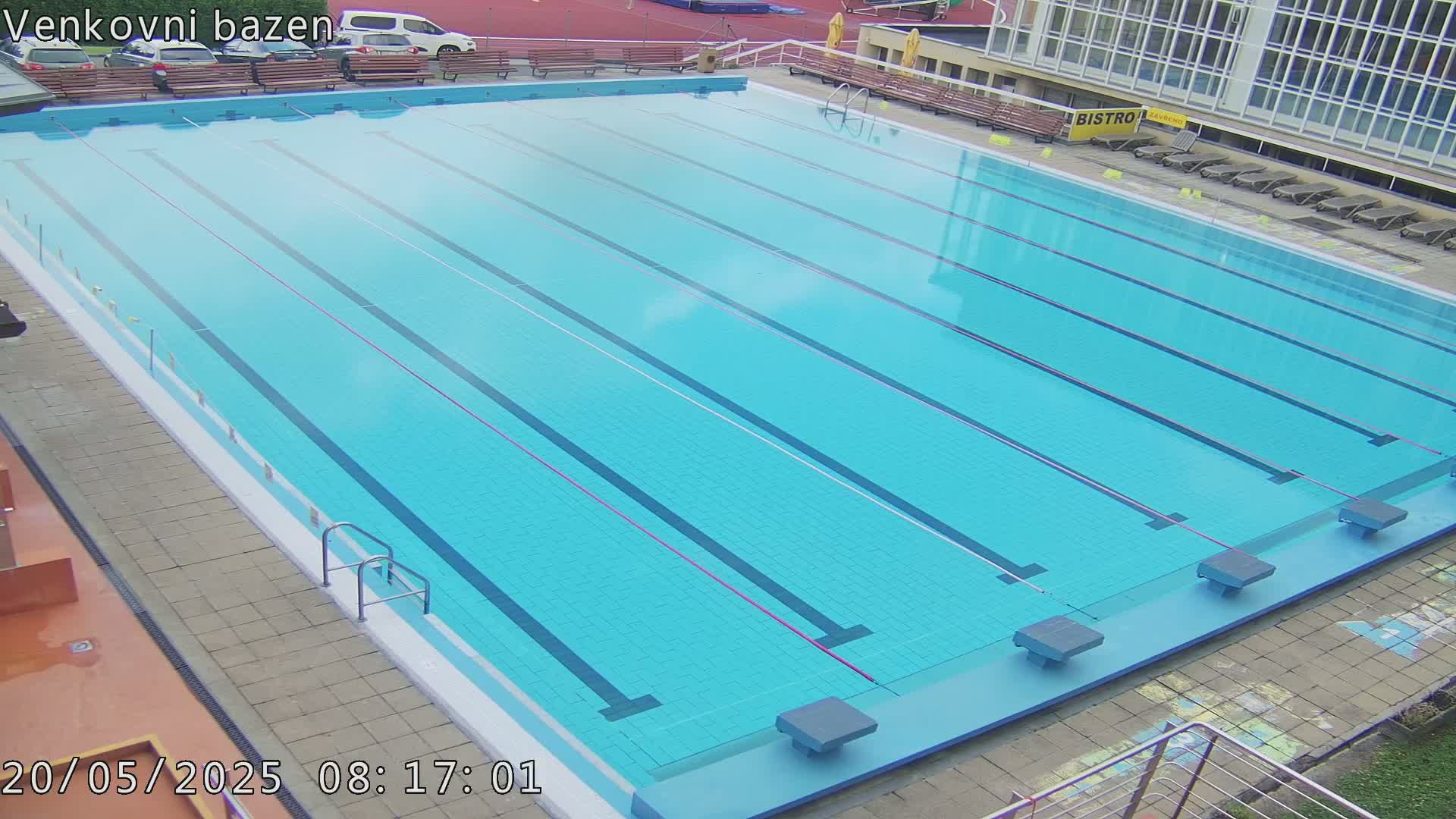 An outdoor lap pool with light blue water and multiple lanes is mostly empty, under a seemingly clear sky.
