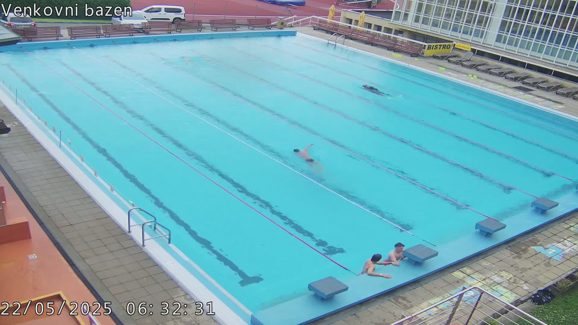 Several people swim in a light blue outdoor swimming pool, with a few others sitting on the edge.