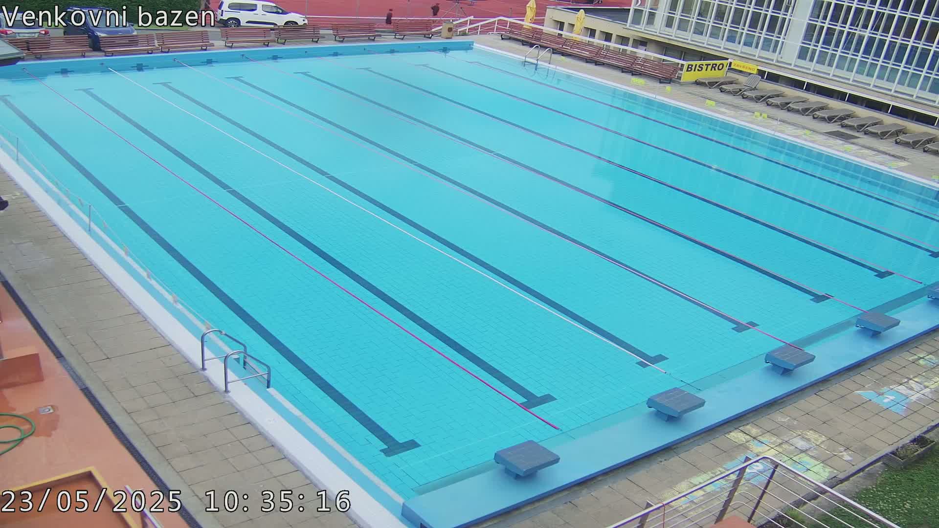 An outdoor swimming pool with multiple lanes is shown, appearing calm and clear under an overcast sky.