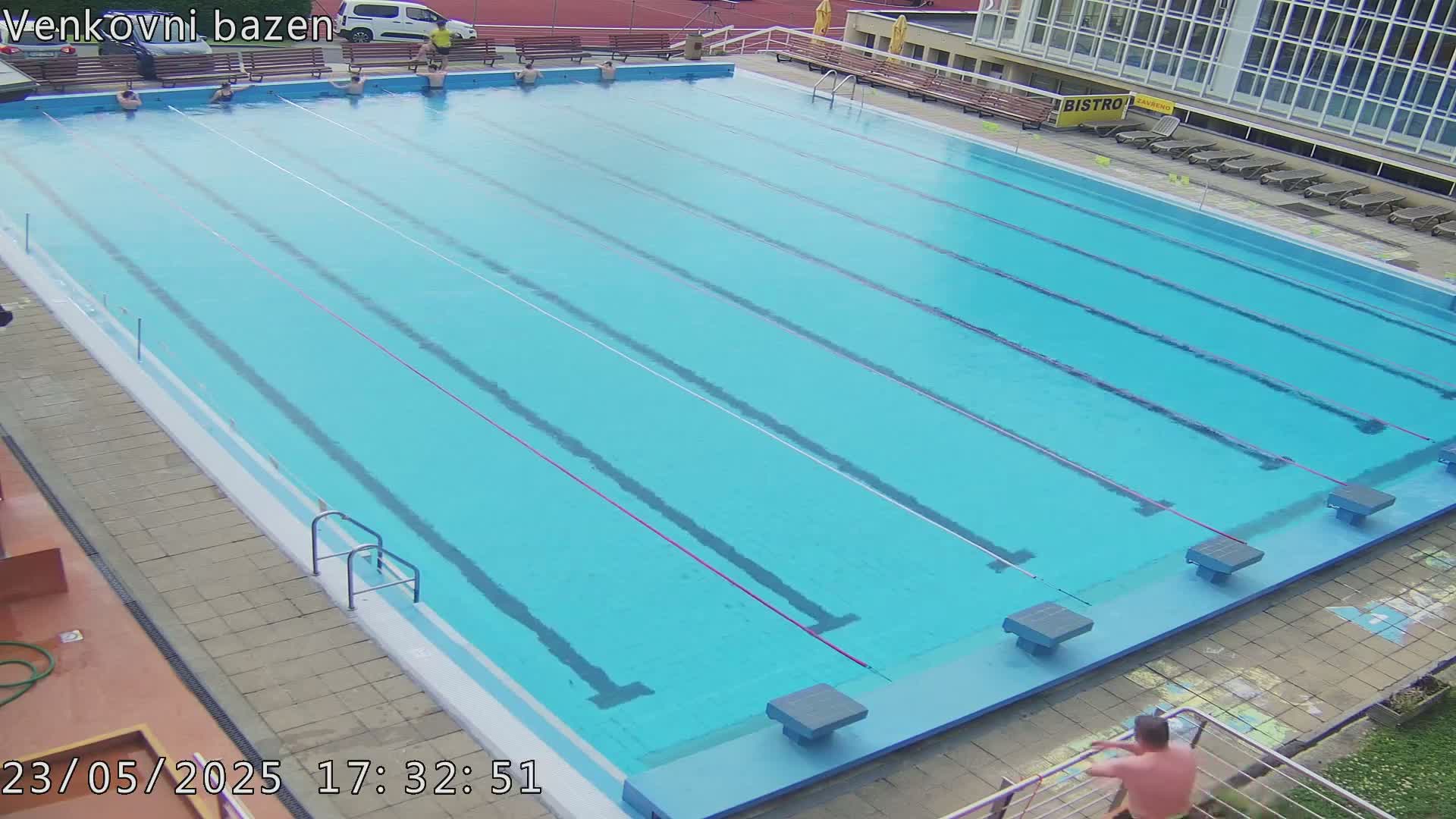An outdoor swimming pool with several lanes and a few people in the water and on the sides, under a mostly clear sky.