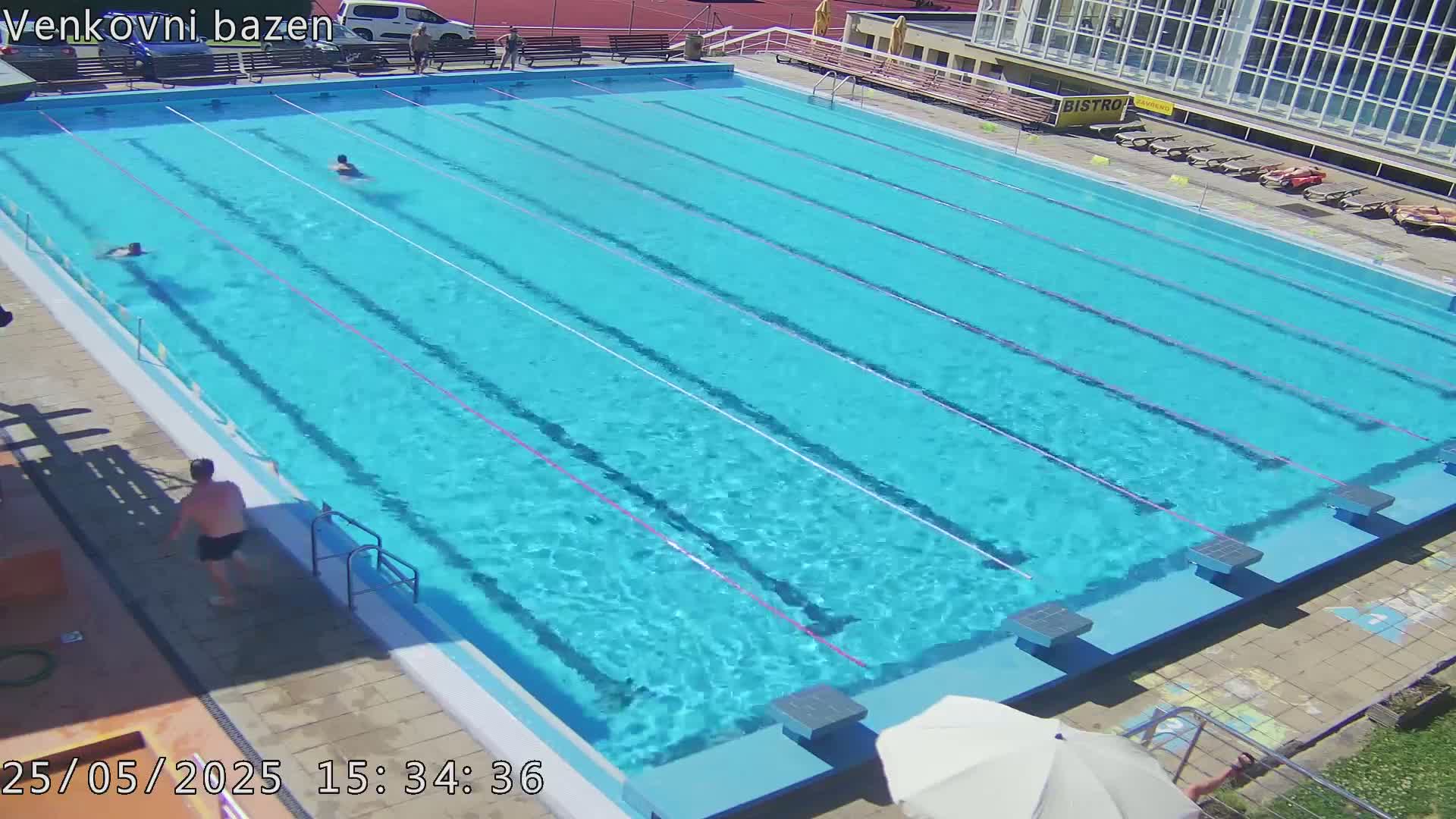 A sunny day at an outdoor swimming pool shows several people swimming in a large, light blue pool with multiple lanes.