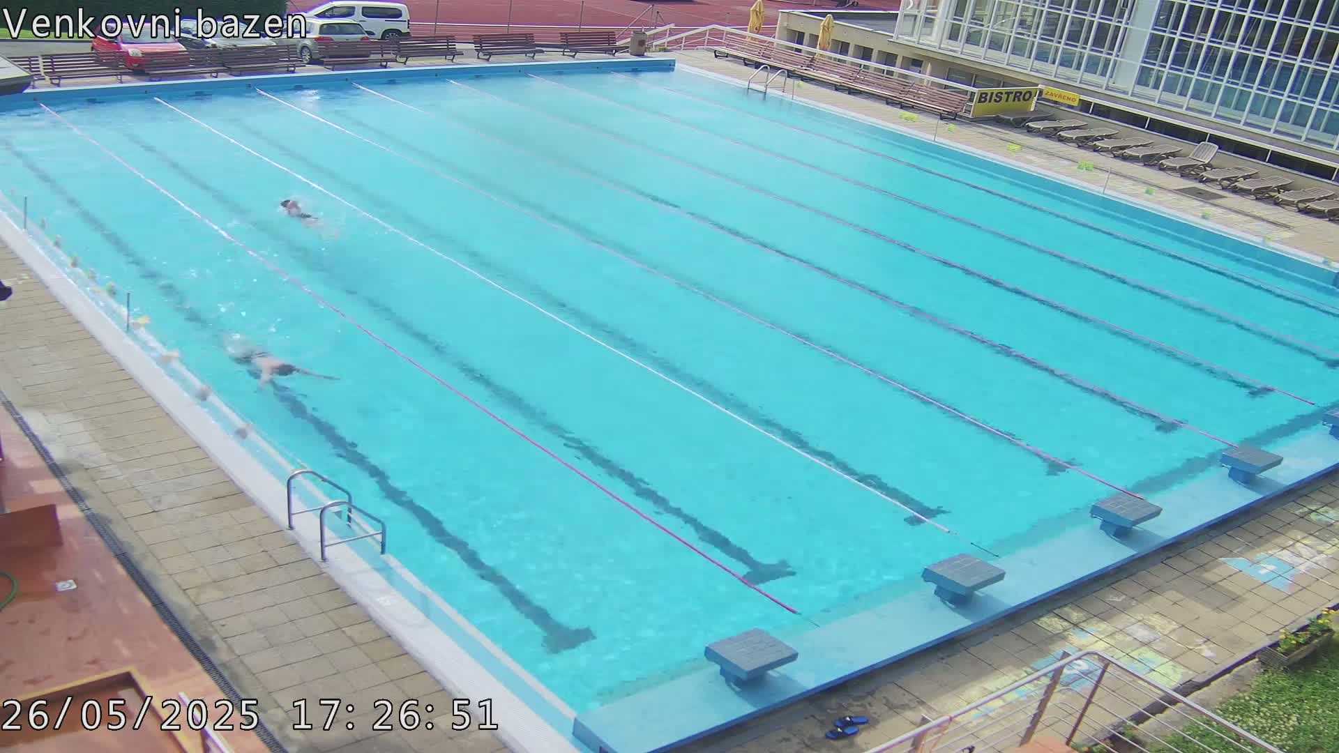 An outdoor swimming pool with several lanes and two people swimming laps under a sunny sky.