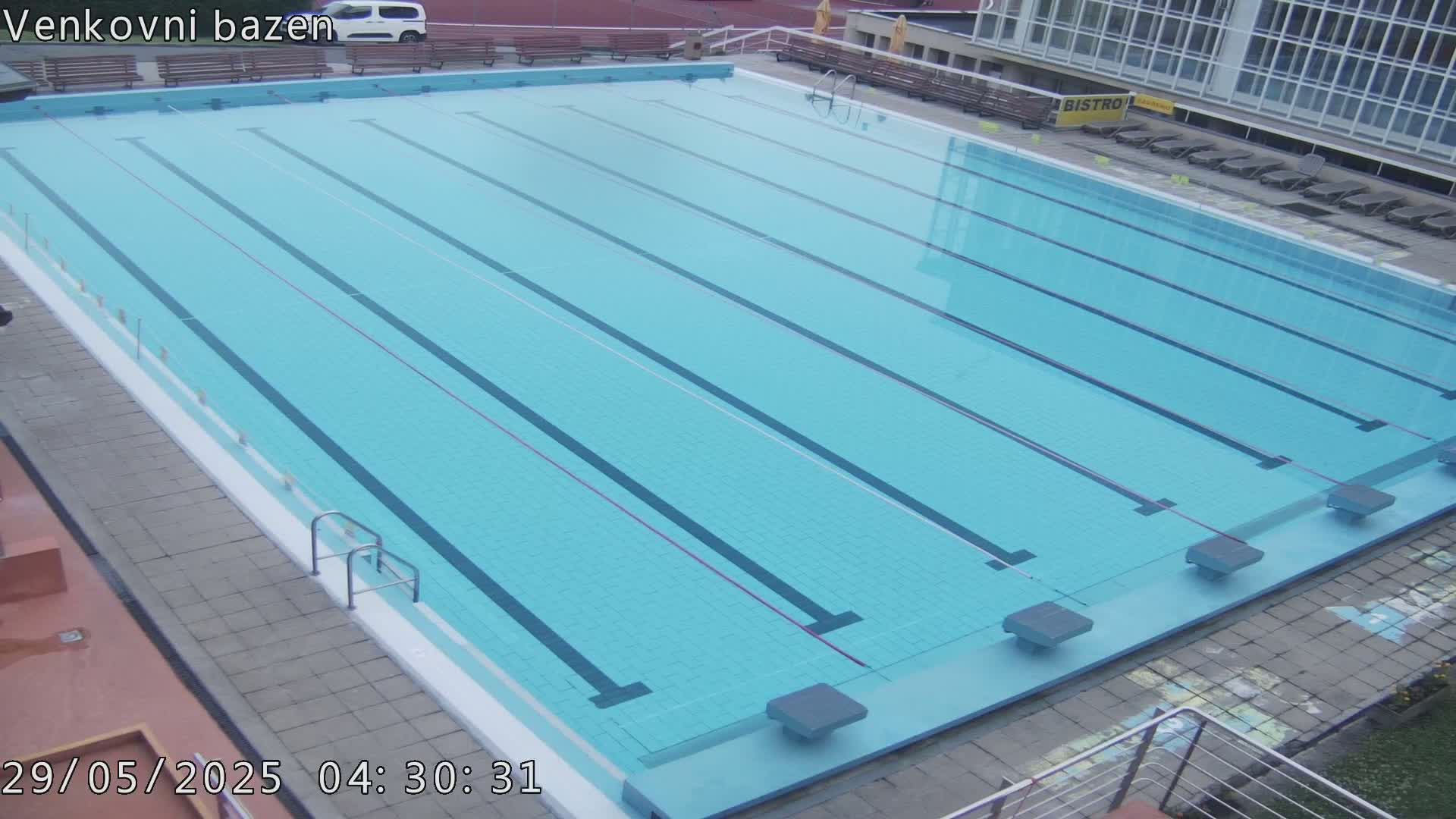 An outdoor, light-blue swimming pool with multiple lanes is empty under an overcast sky.