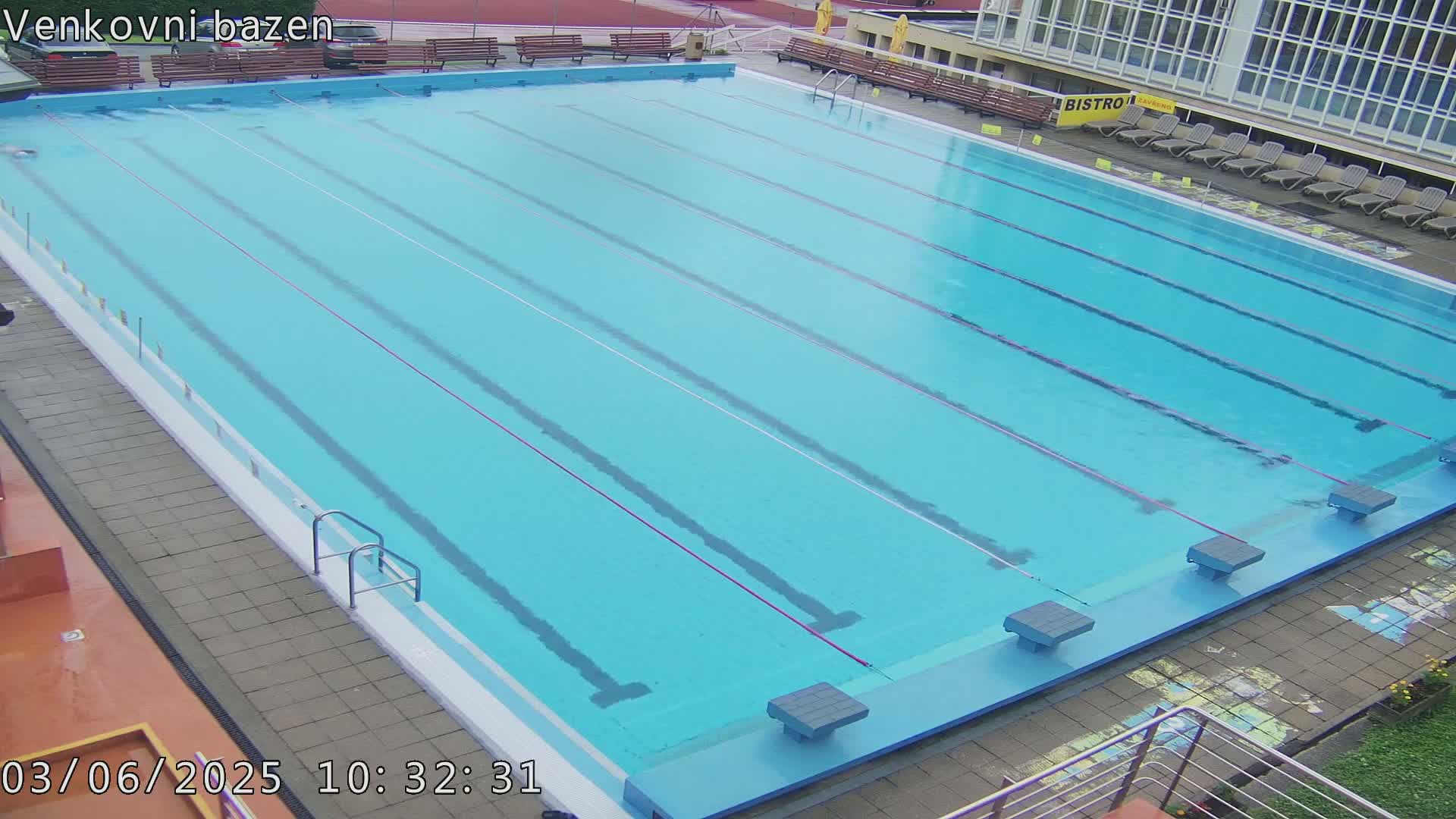 An outdoor swimming pool with lane dividers is mostly empty, under a cloudy sky.