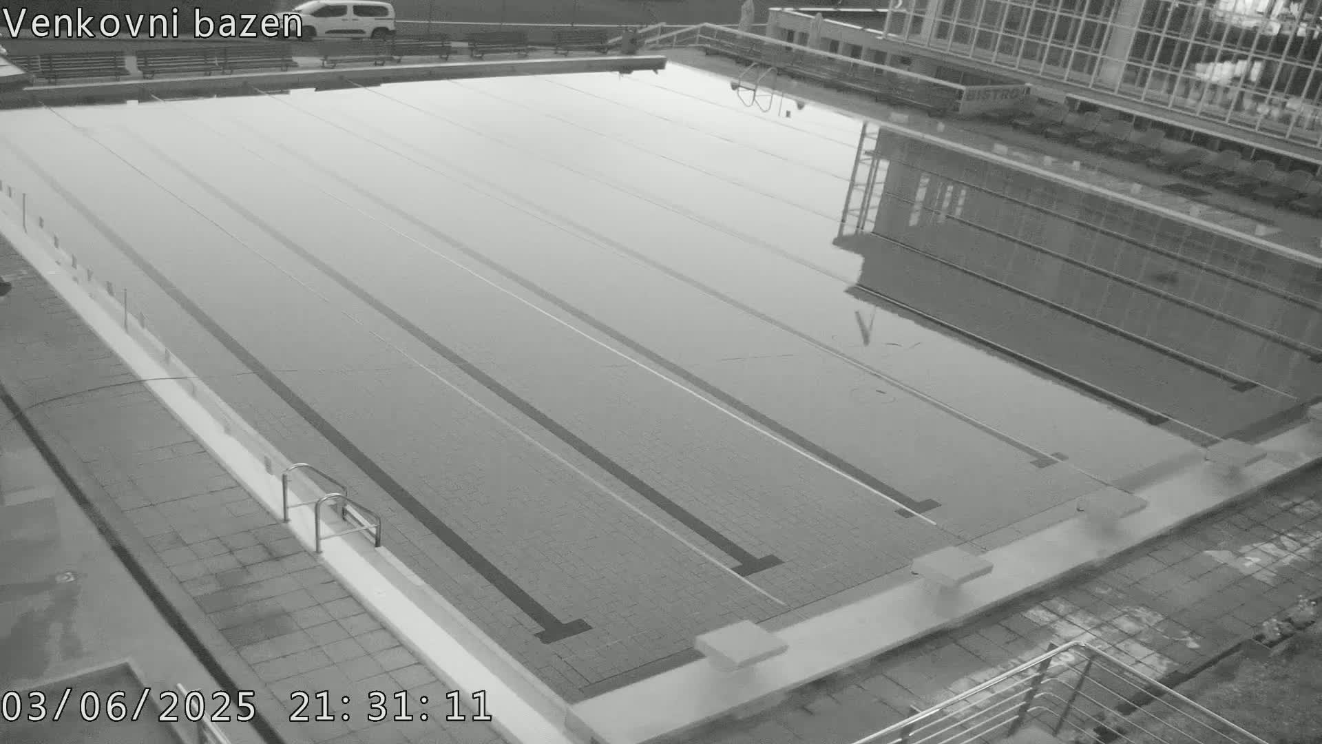 An empty outdoor swimming pool is seen at night, reflecting the surrounding buildings in its still water.