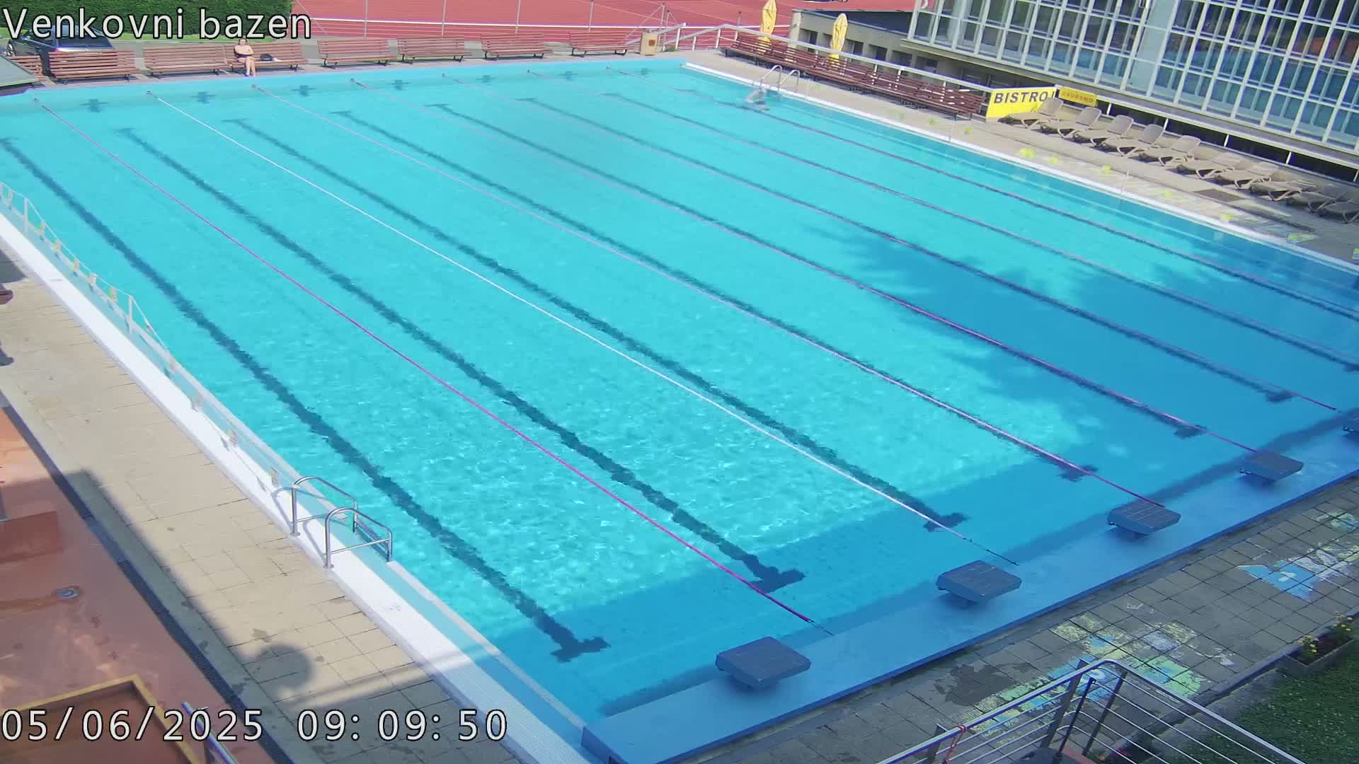 An outdoor swimming pool with multiple lanes is mostly empty, under sunny conditions.