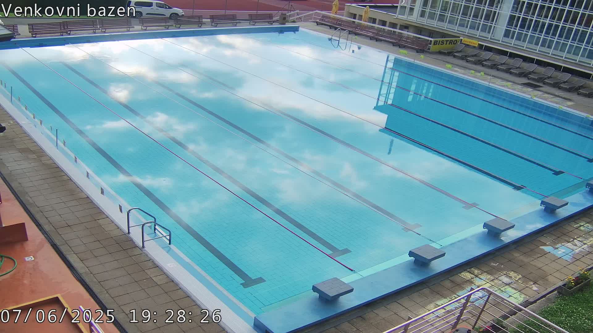 An outdoor swimming pool with lane dividers reflects a partly cloudy sky.