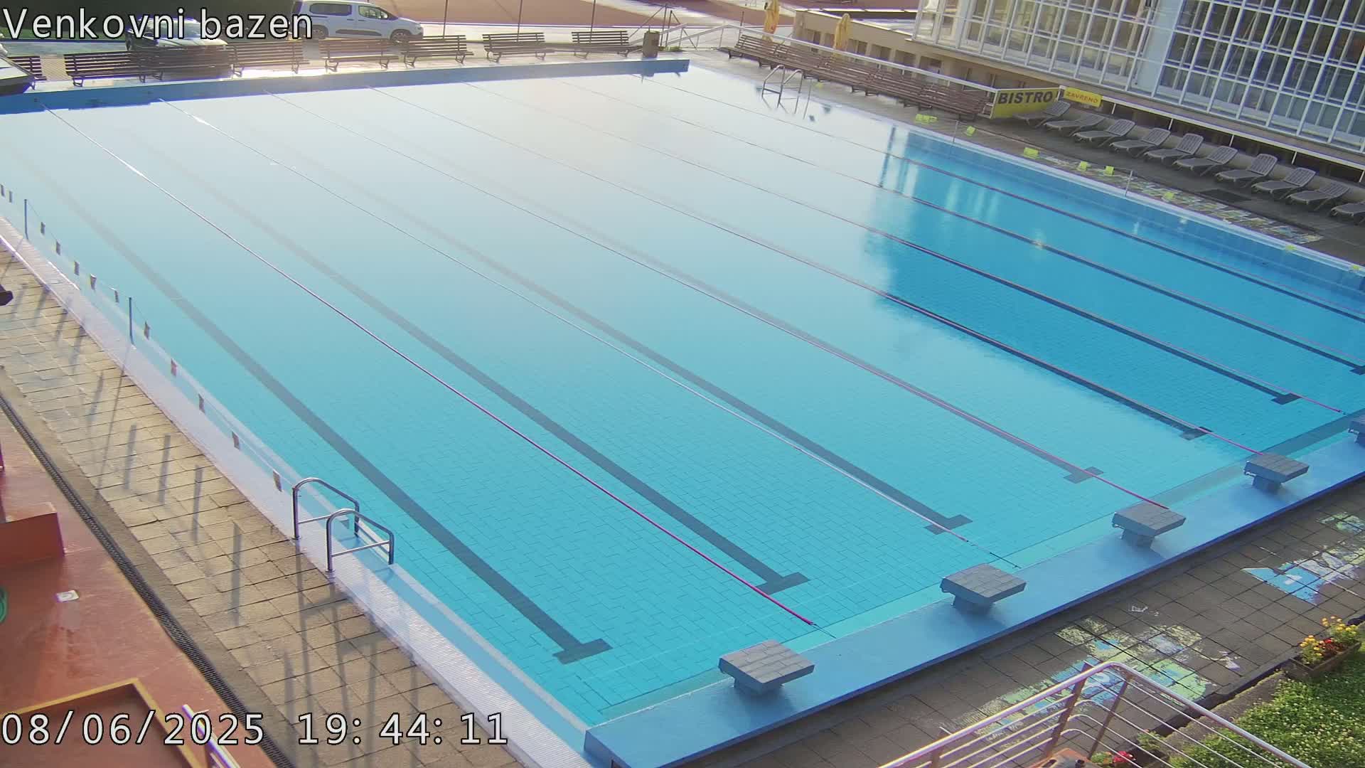 An outdoor swimming pool with multiple lanes is mostly empty, under sunny conditions.