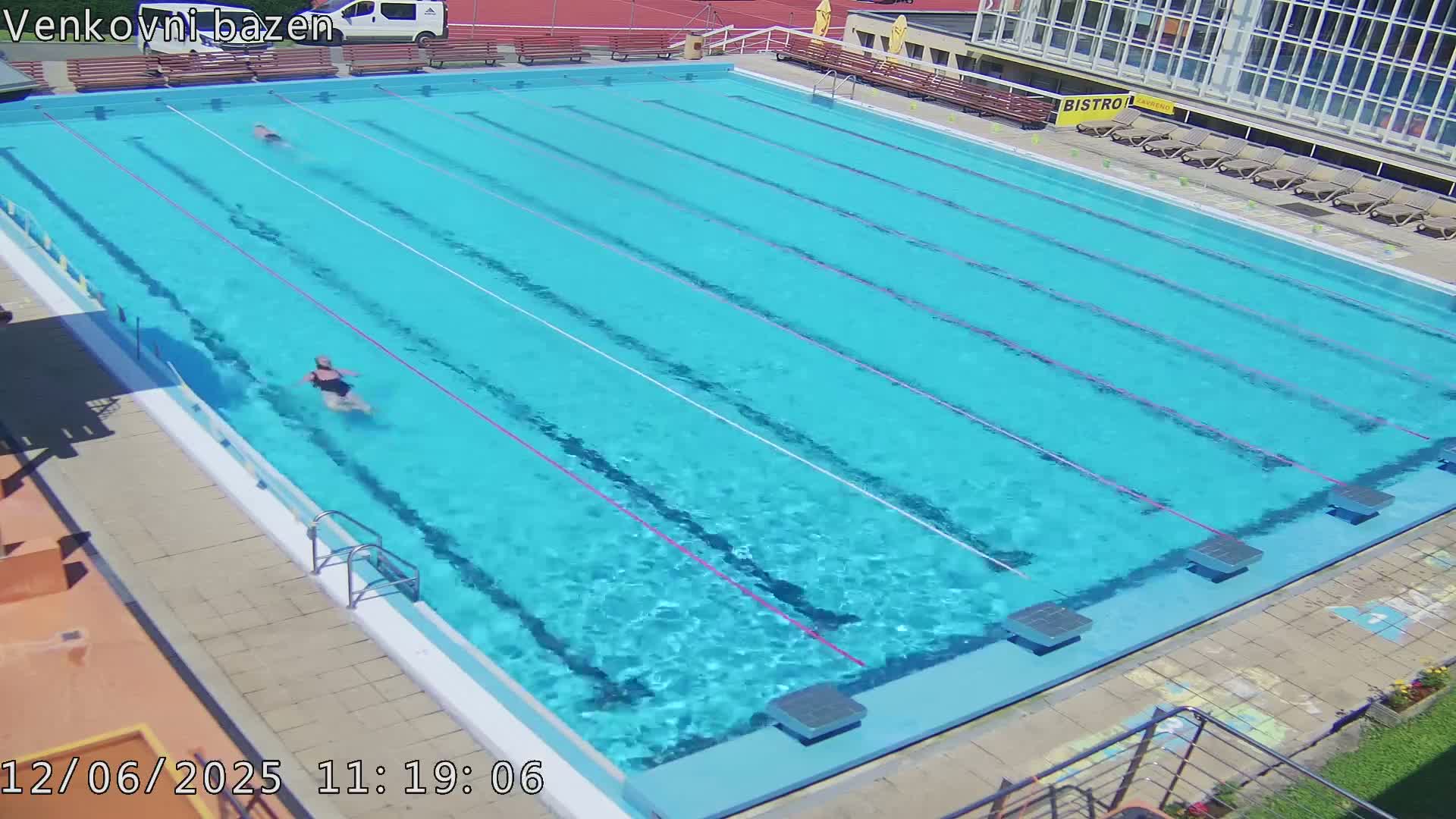 An outdoor swimming pool with several lanes and a few people swimming in it on a sunny day.