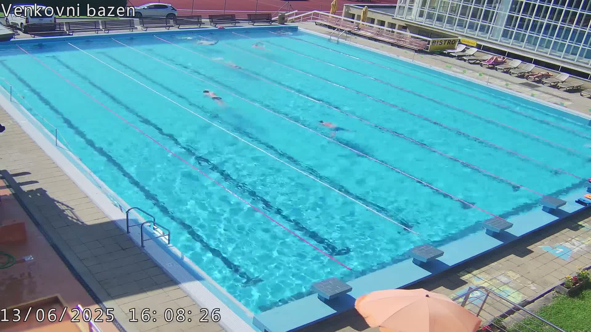 A large outdoor swimming pool with several people swimming laps in bright, sunny weather.