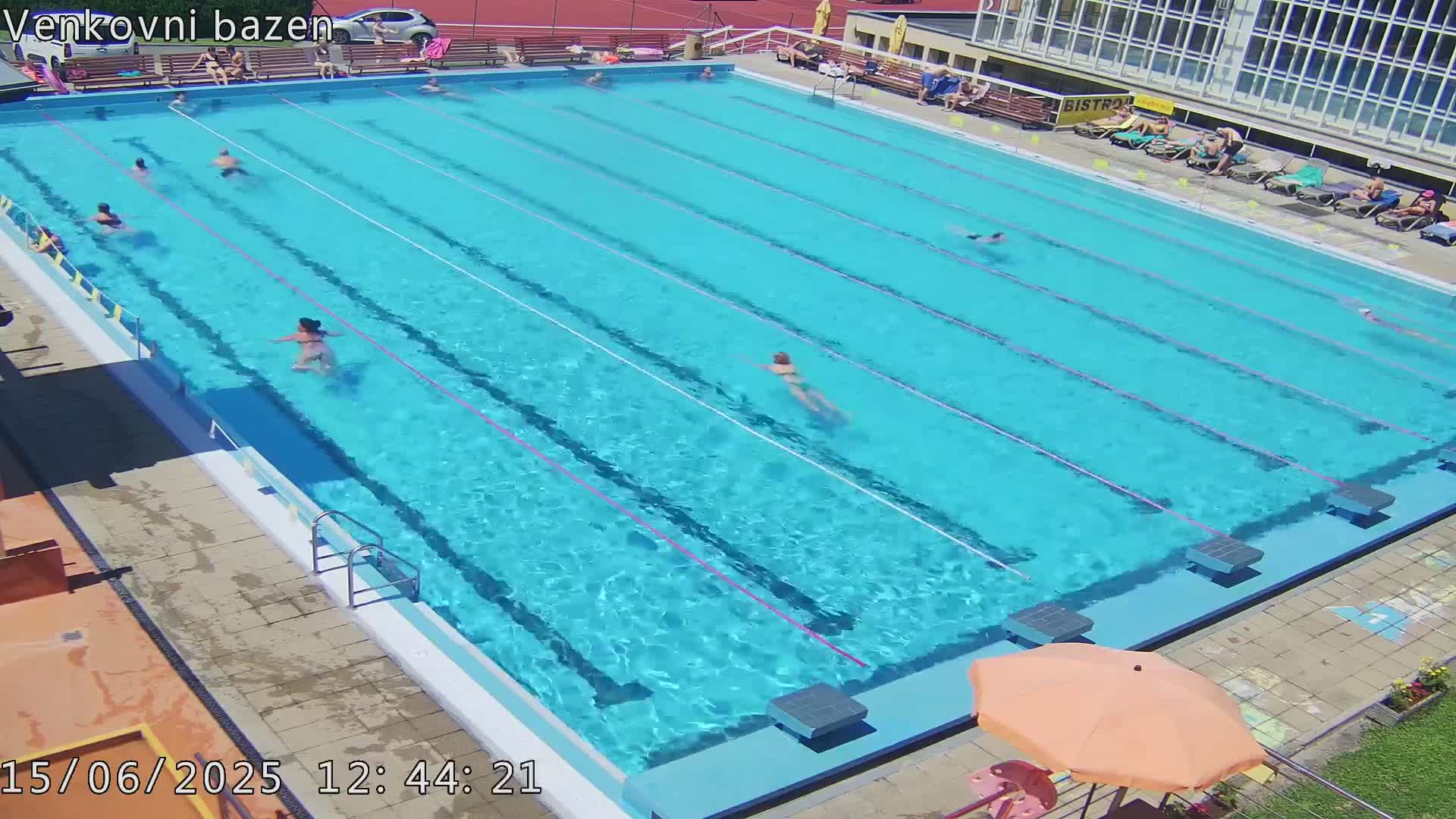 A large outdoor swimming pool with multiple people swimming and others relaxing on the surrounding deck on a sunny day.