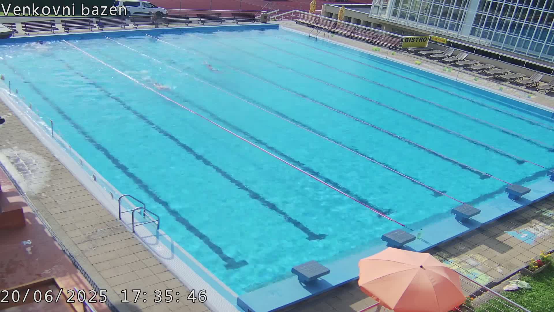 An outdoor swimming pool with several lanes, a few people swimming, and an orange umbrella nearby on a sunny day.