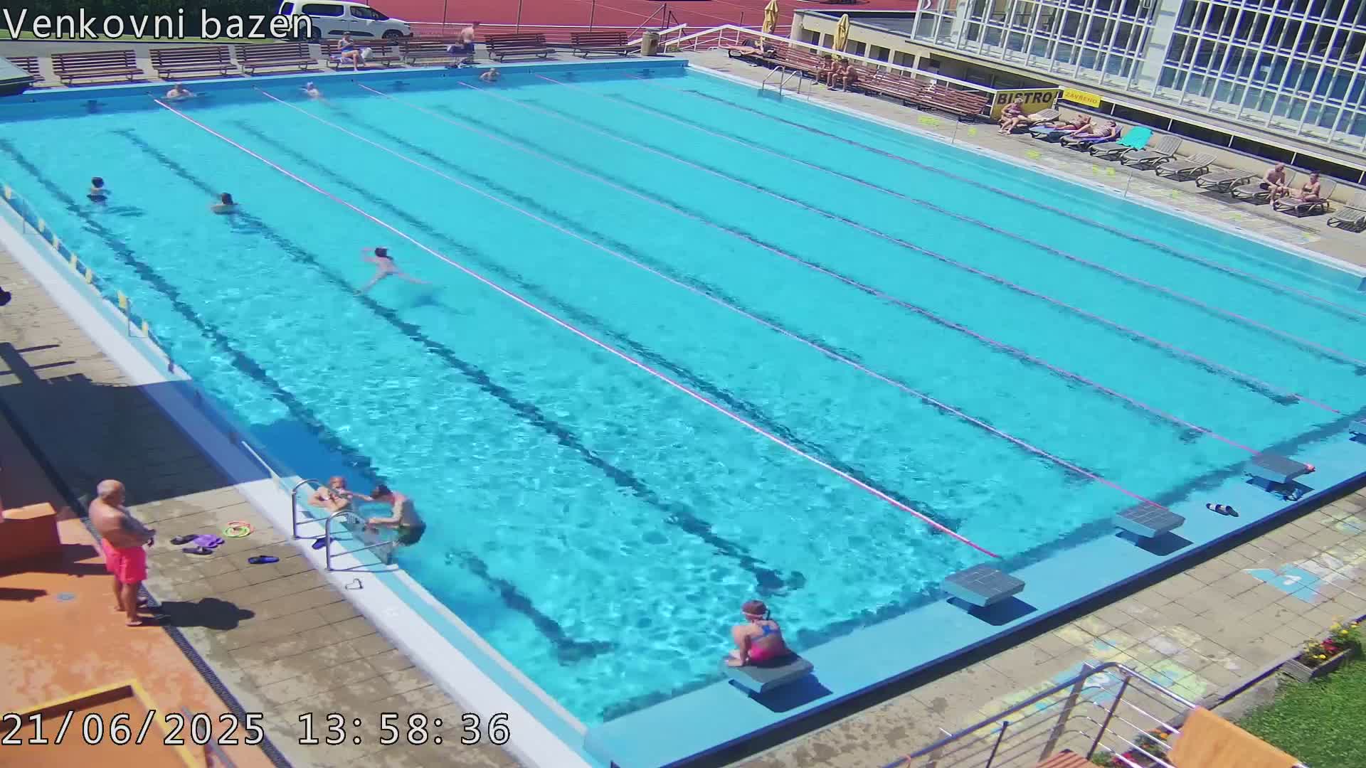 A large outdoor swimming pool with several people swimming and relaxing on the sunny pool deck.