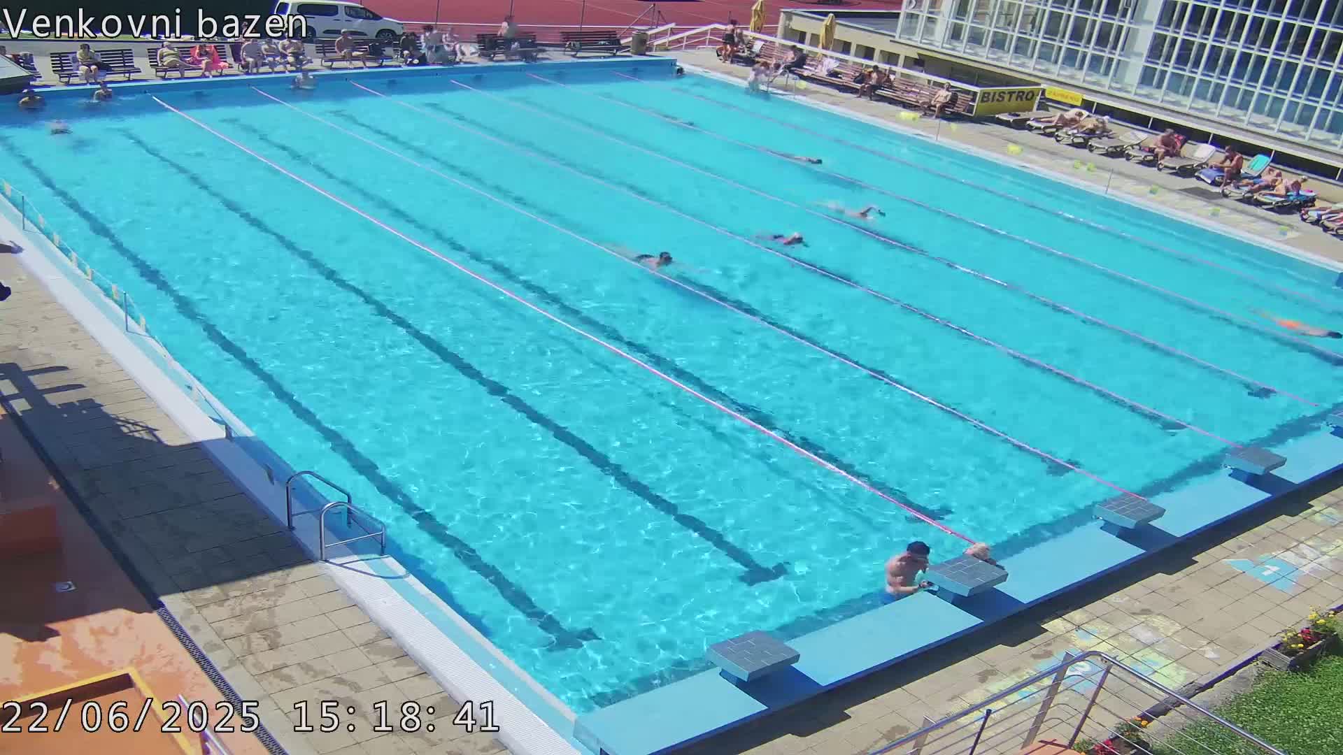 A large outdoor swimming pool with several people swimming and sunbathing on a sunny day.