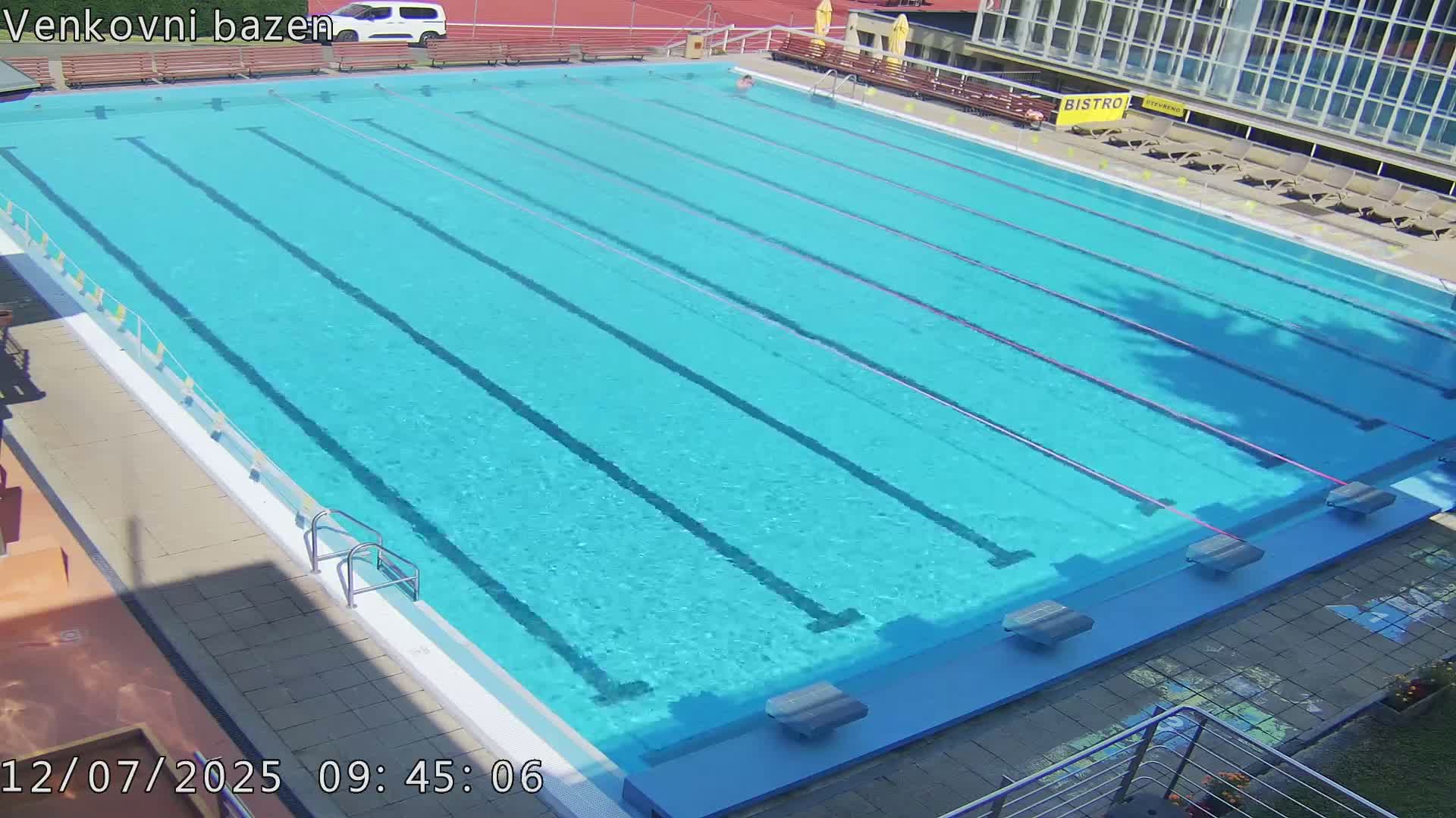 A sunny day at an outdoor swimming pool shows a person swimming laps in a long, rectangular pool with multiple lanes.