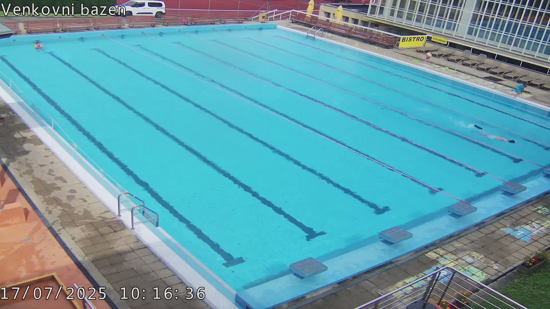 An outdoor swimming pool with multiple lanes has a few people swimming in it on a sunny day.