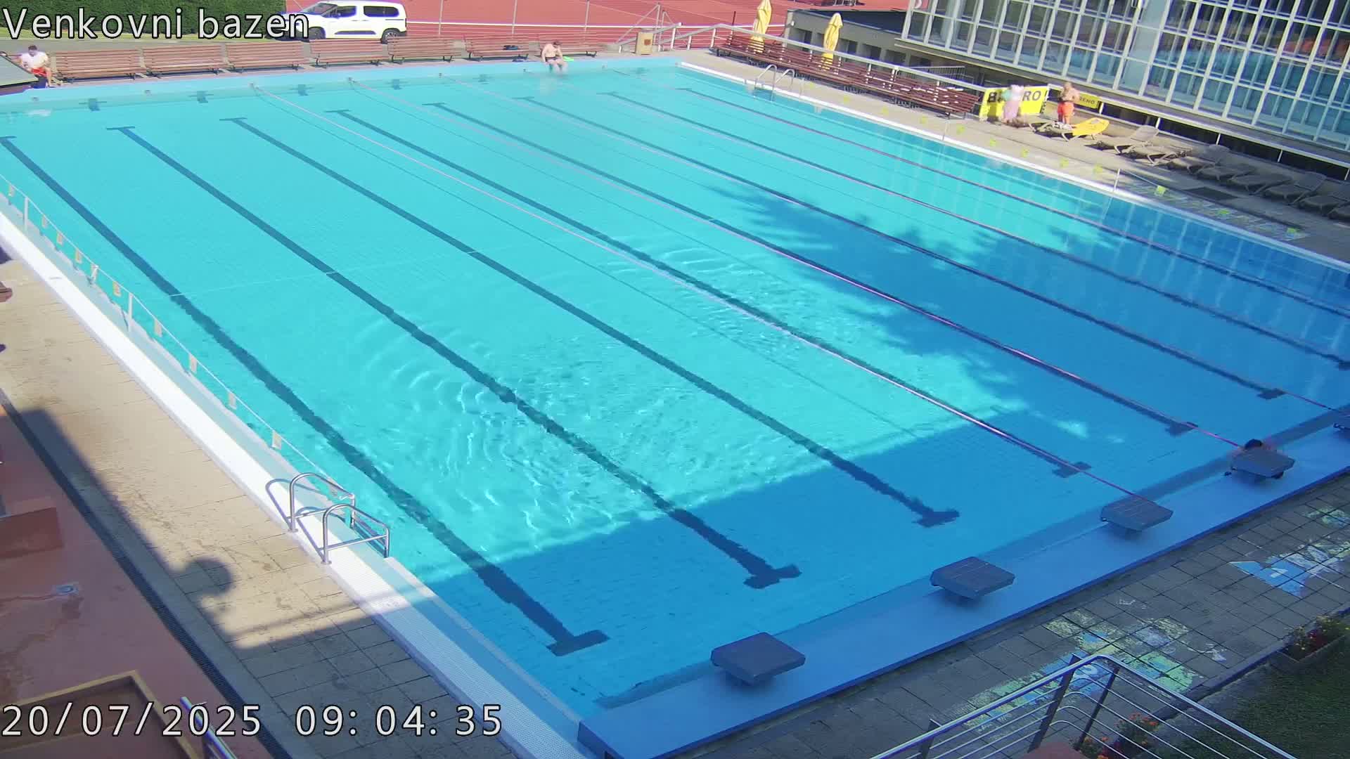 A mostly empty, sunny outdoor swimming pool with multiple lanes and a few people visible near the edges.