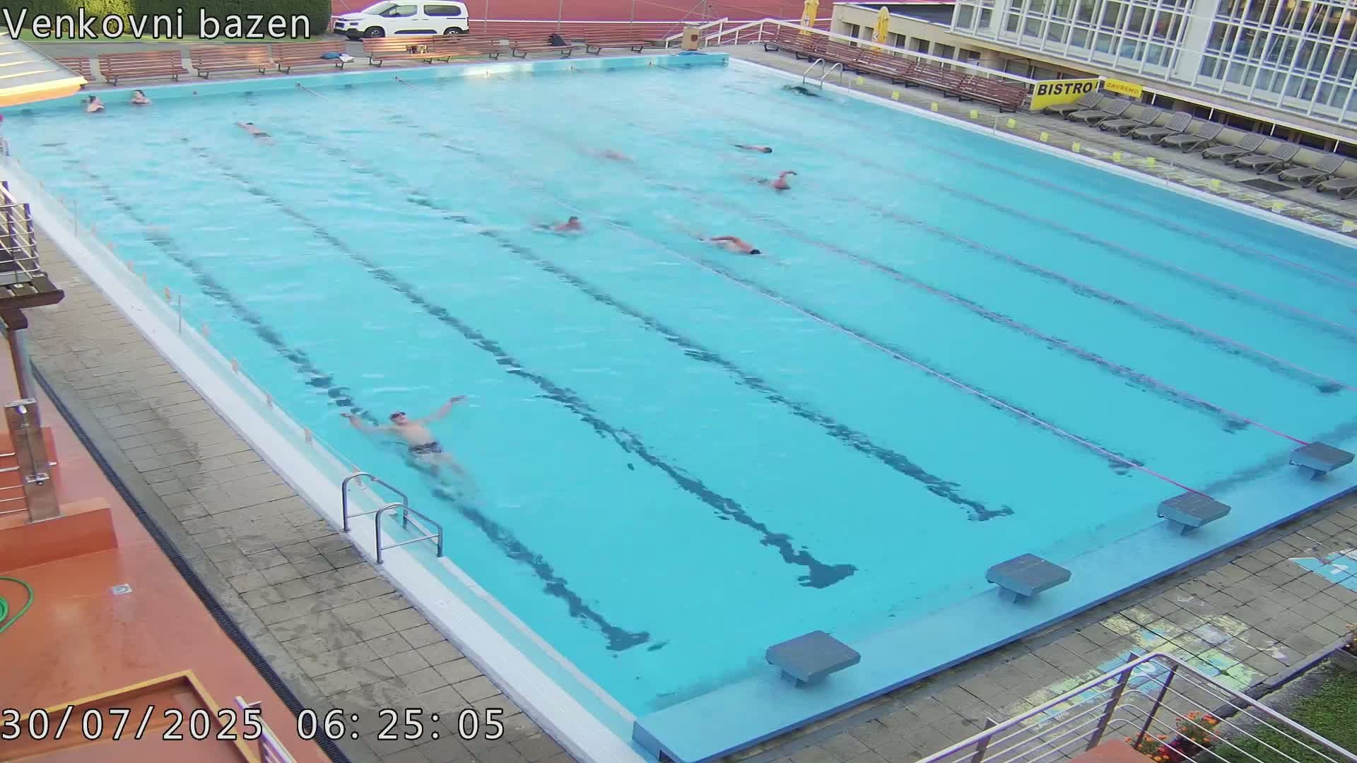 A mostly empty, sunny outdoor swimming pool with multiple lanes and a few people visible near the edges.