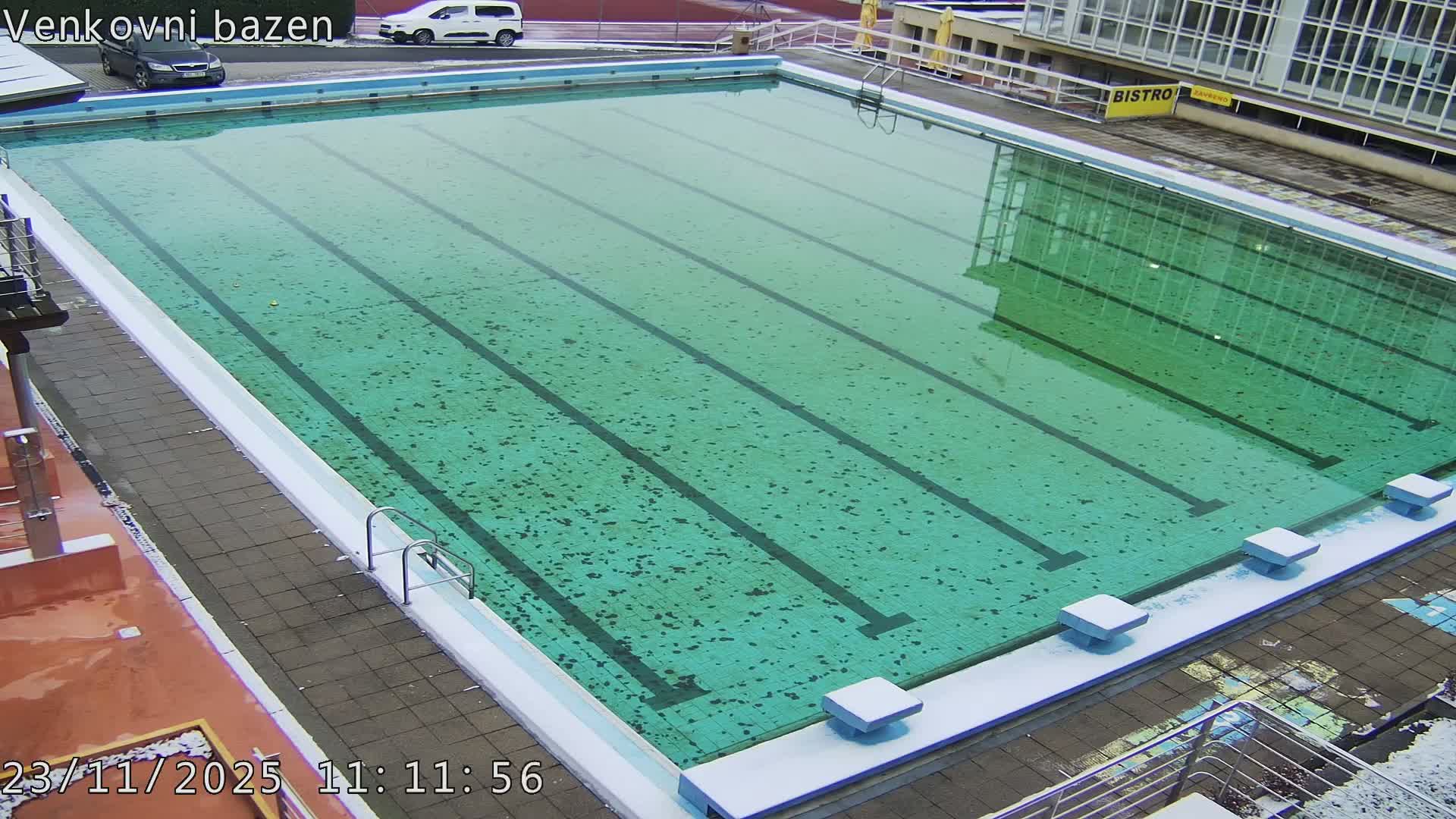 An outdoor swimming pool with murky green water and dark lane lines is visible from above, surrounded by snow and ice on the paved edges, under cold and likely overcast weather.