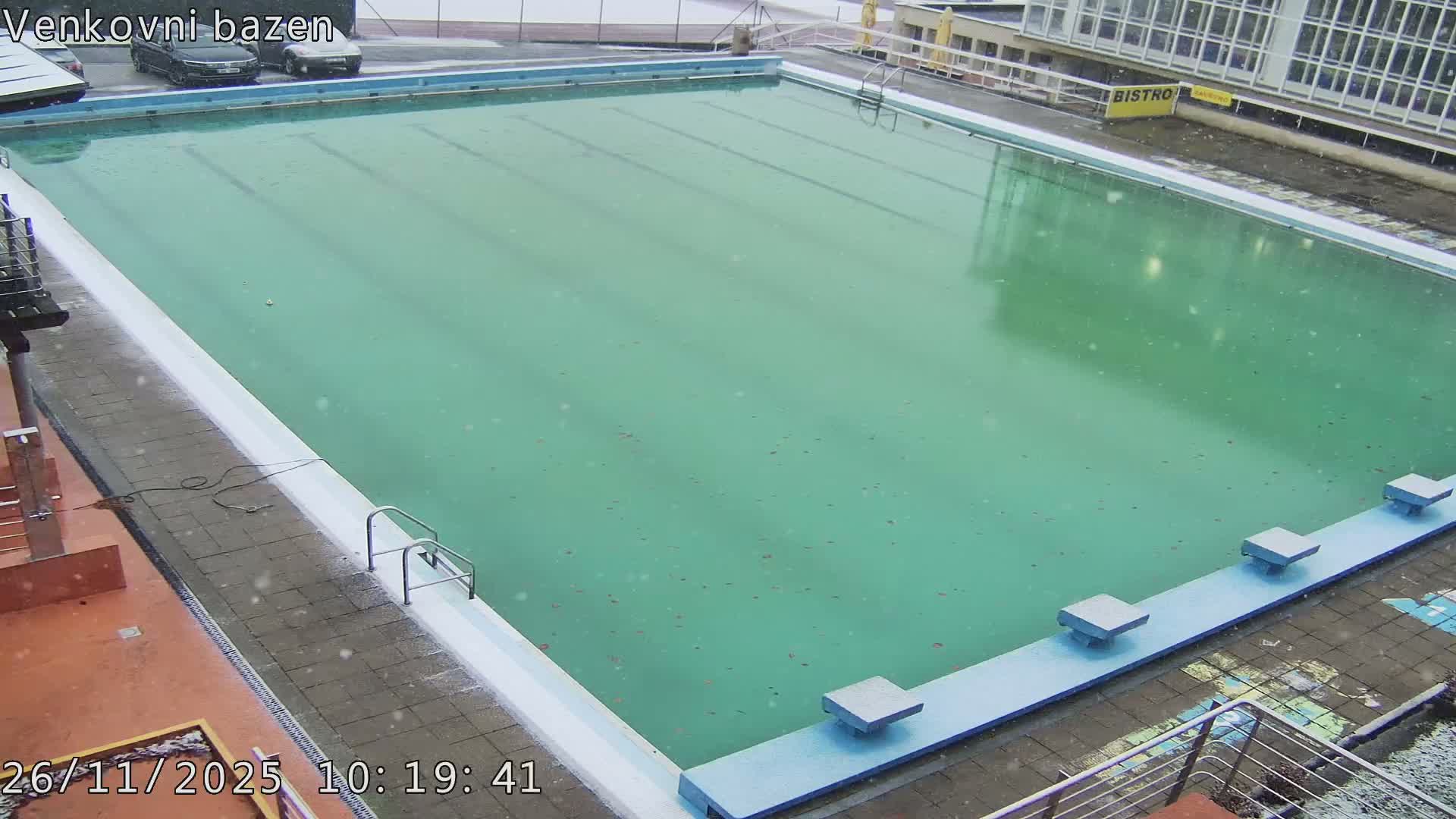 An outdoor swimming pool with teal-colored water and starting blocks is seen under a light snowfall, with paved areas and buildings in the background.